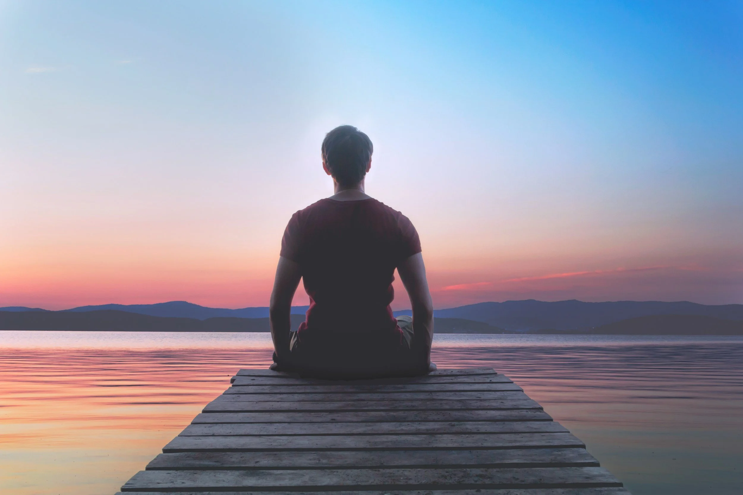 Person sitting on a pier by a lake during sunset, facing the water with mountains in the distance.