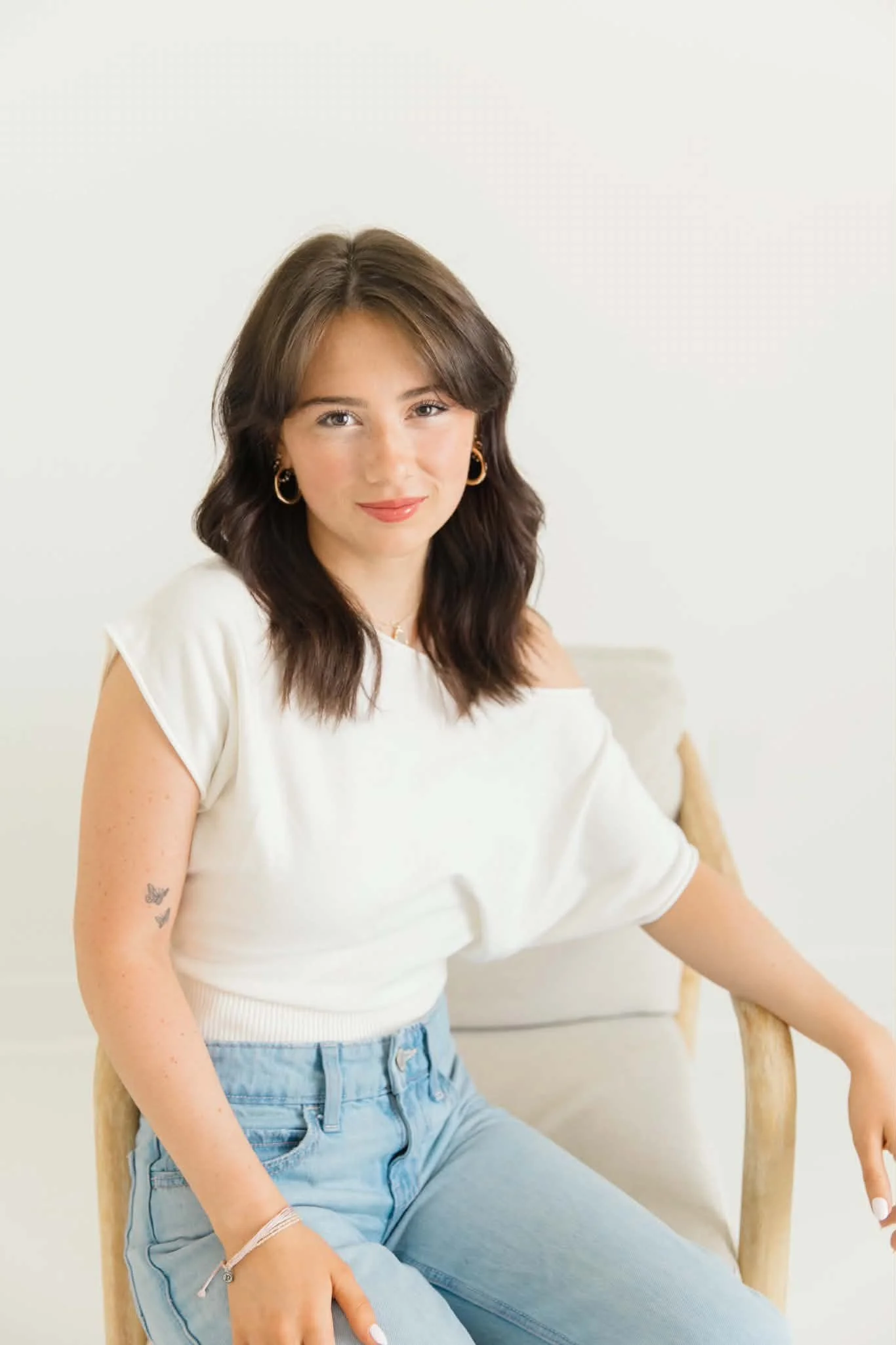 A young woman with shoulder-length brown hair, wearing gold hoop earrings and a white top. She is sitting on a wooden chair against a plain light background, smiling softly, with her left arm resting on the chair's armrest.