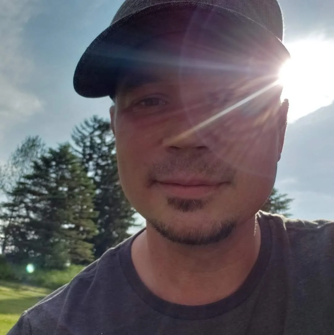 A man wearing a black cap and a gray t-shirt is outdoors on a sunny day with trees in the background. The sun is shining directly into the camera, creating a lens flare over his face.