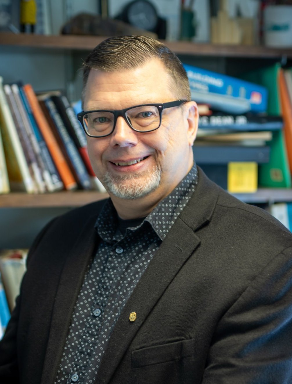 Man wearing glasses and a black blazer, smiling, standing in front of a bookshelf.