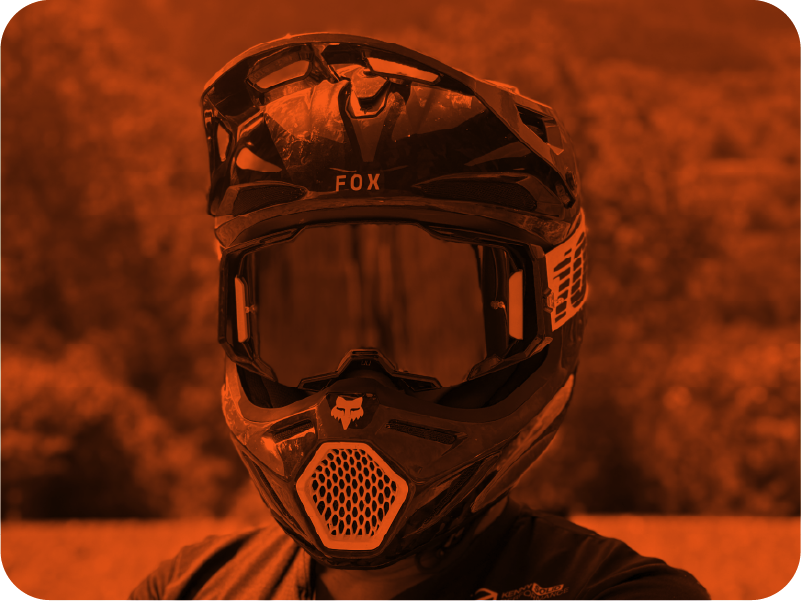 Un casque de moto tout noir avec une visière sombre, posé sur la tête d'une personne dont le visage n'est pas visible, avec un arrière-plan flou. L'image a une teinte orangée.