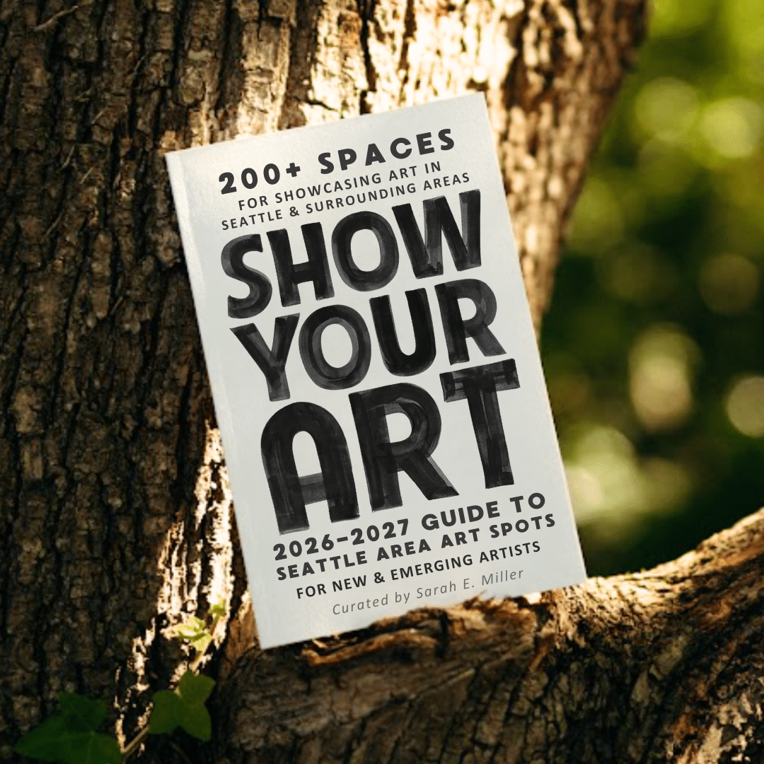 A book rests in the crook of a tree; its cover reads "200+ spaces for showcasing art in Seattle & surrounding areas. SHOW YOUR ART 2026-2027 guide to Seattle area art spots for new & emerging artists. Curated by Sarah E. Miller."