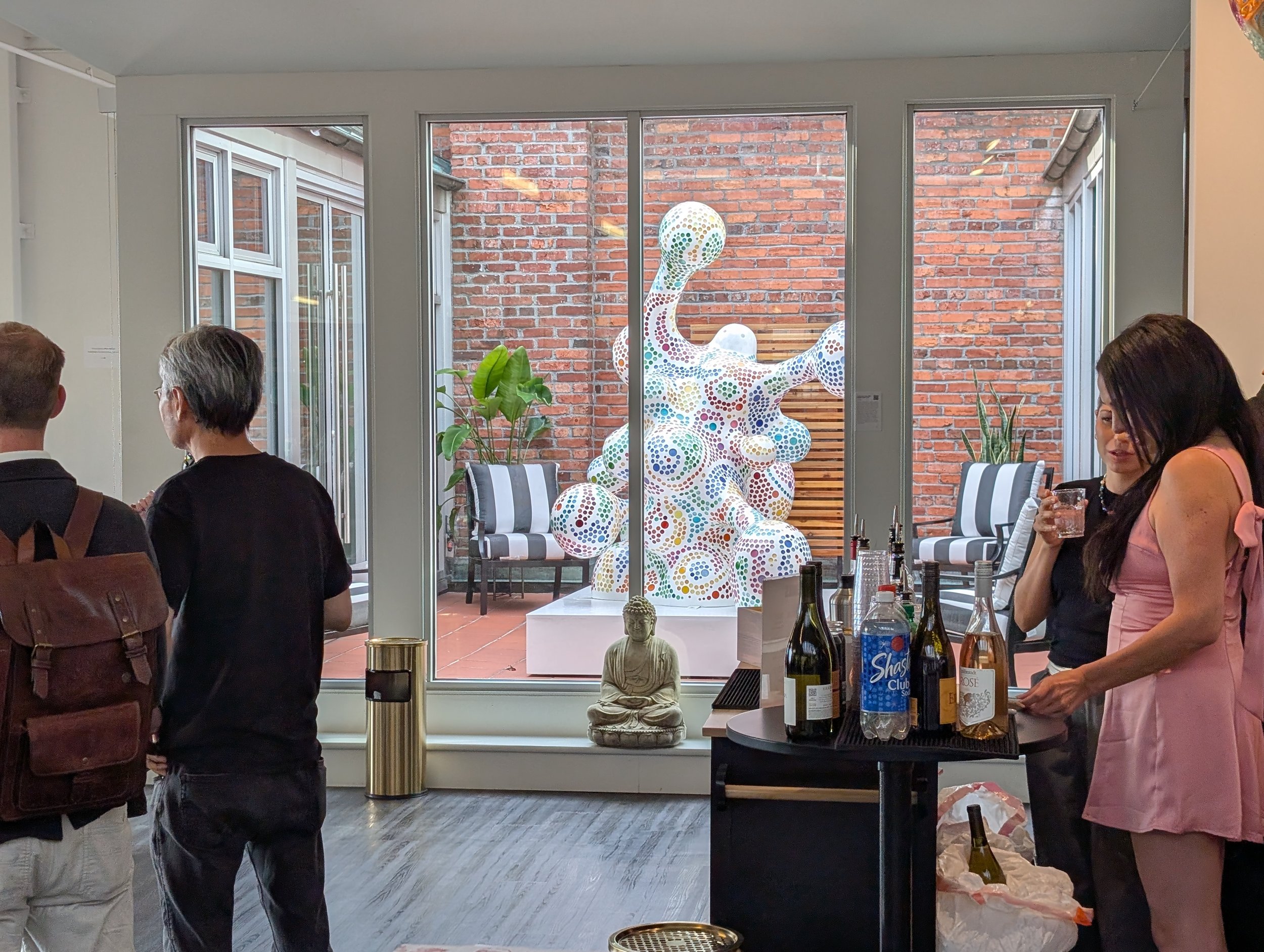 Gallery visitors at ArtLove Salon mingle with beverages in the foreground; in the background, the gallery's courtyard is visible, with a large white abstract sculpture at its center, covered in colorful polka dots. Its shape is like a cartoon amoeba.