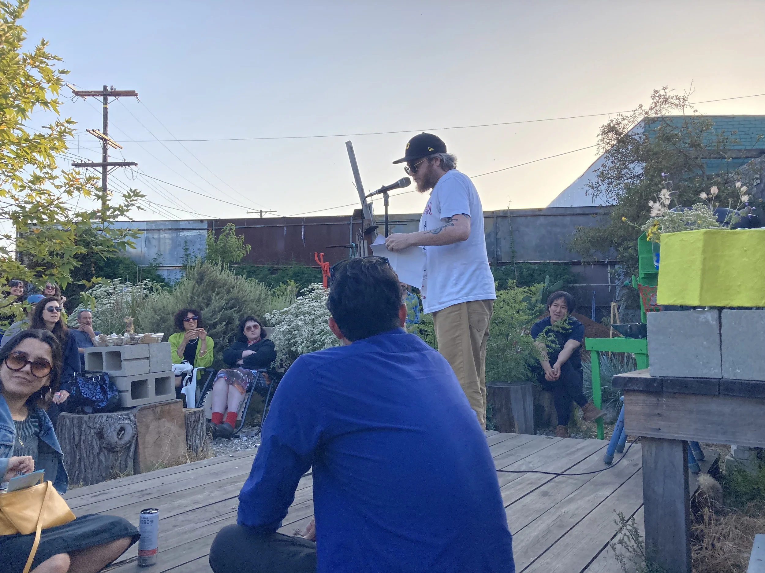 Ed Steck, a Pittsburgh poet, reads from loose leaves of paper on a wooden stage in an urban garden at dusk. Wearing a ballcap, sunglasses, a grey tee and tan pants, he leans towards a mic to address the crowd around him.