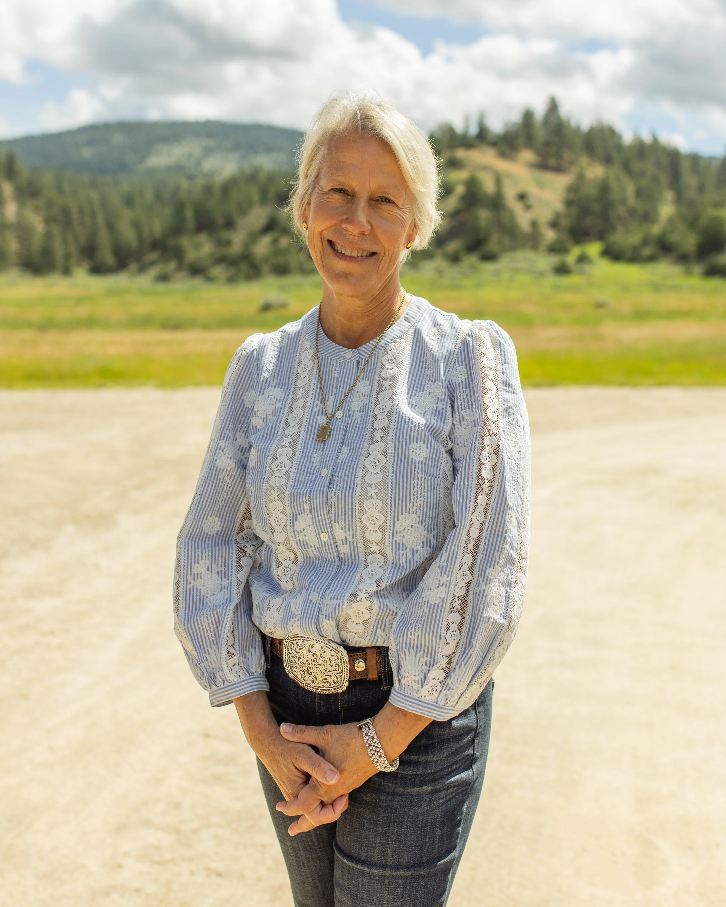 An older woman with short gray hair smiling outdoors, wearing a light blue and white striped blouse with lace patterns, dark jeans with a fancy belt, a wristwatch, and a necklace, standing in a natural landscape with trees, hills, and a partly cloudy sky.
