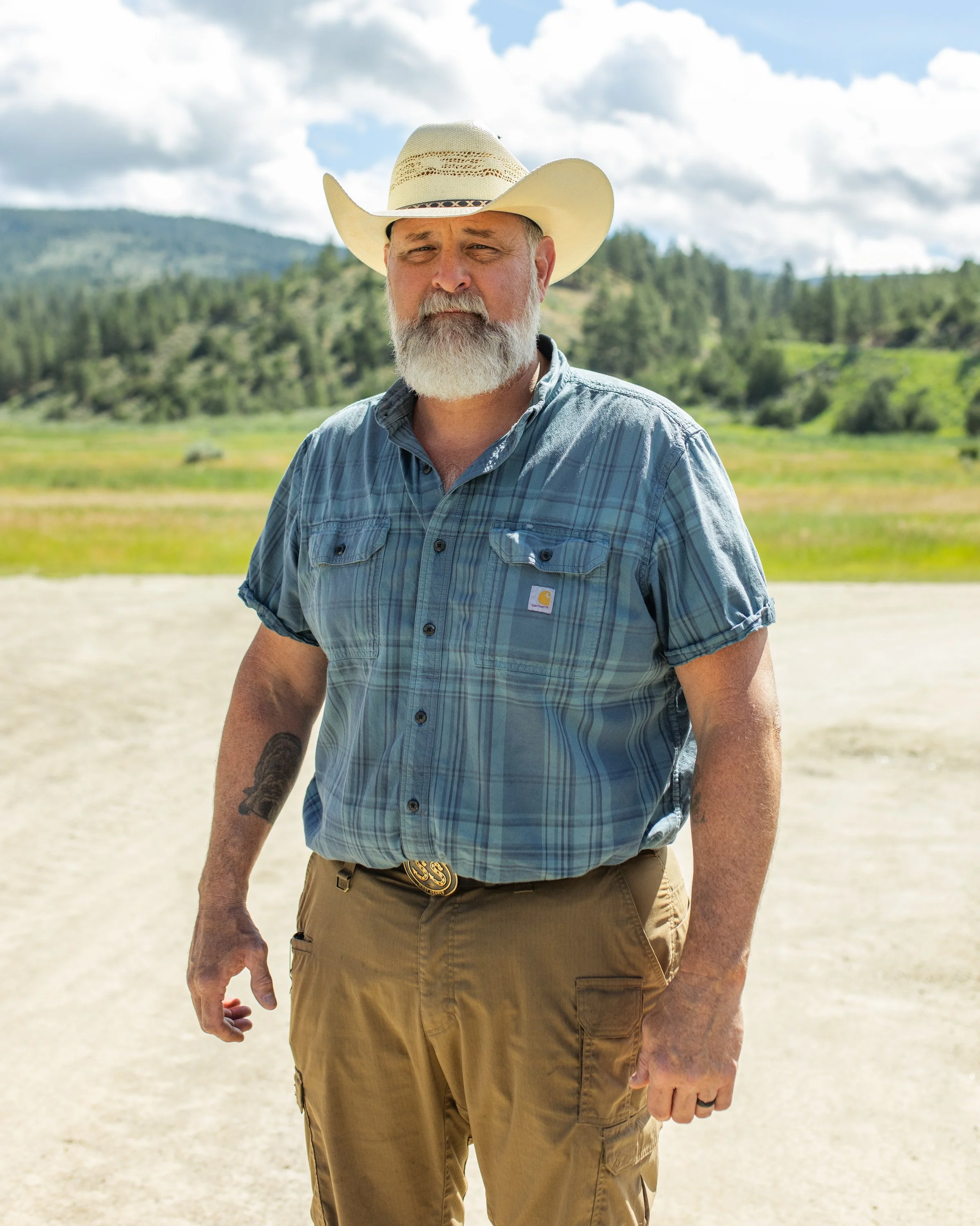 A bearded man in a cowboy hat and blue plaid shirt standing outdoors in a rural area with mountains and trees in the background.