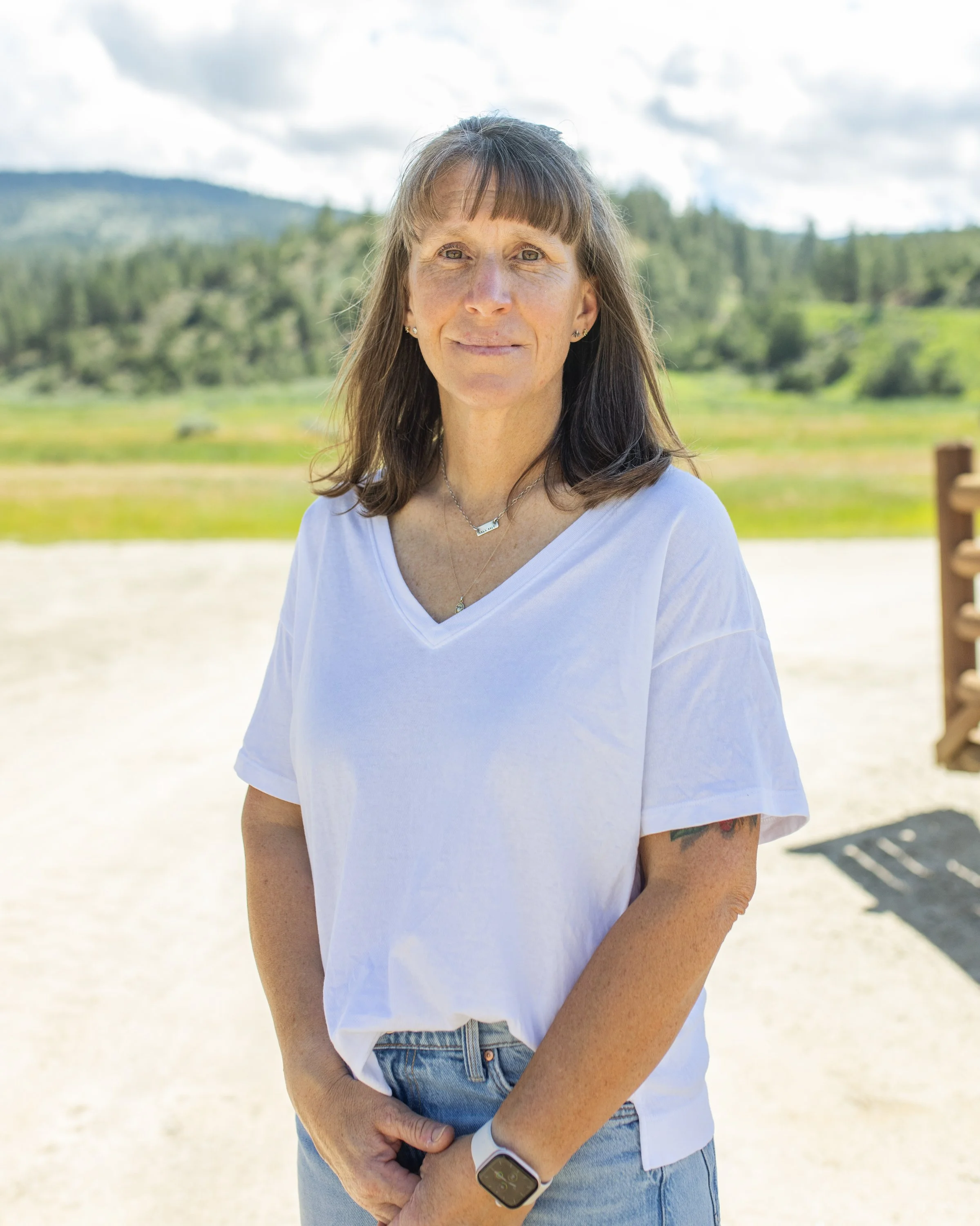 A woman standing outdoors with mountains and greenery in the background, wearing a white t-shirt and jeans.