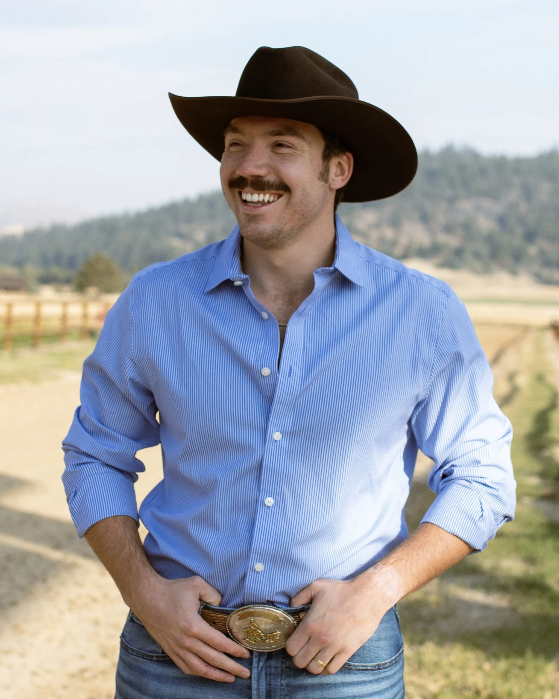 A man wearing a brown cowboy hat and a blue button-up shirt, smiling outdoors with mountains in the background.