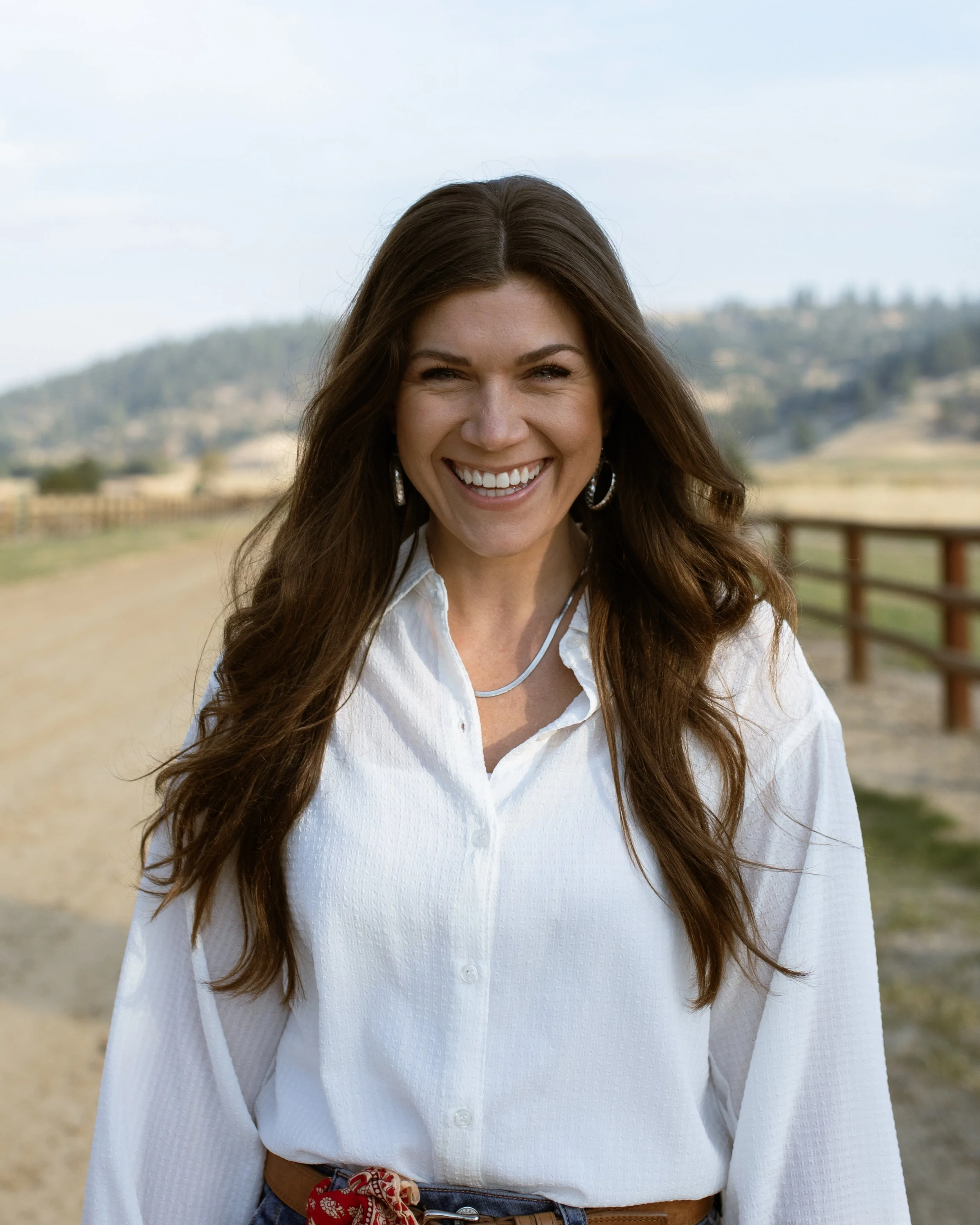 Close-up of a woman smiling outdoors, wearing a white button-up shirt, brown belt with a red bandana, and large earrings, with a rural landscape in the background.
