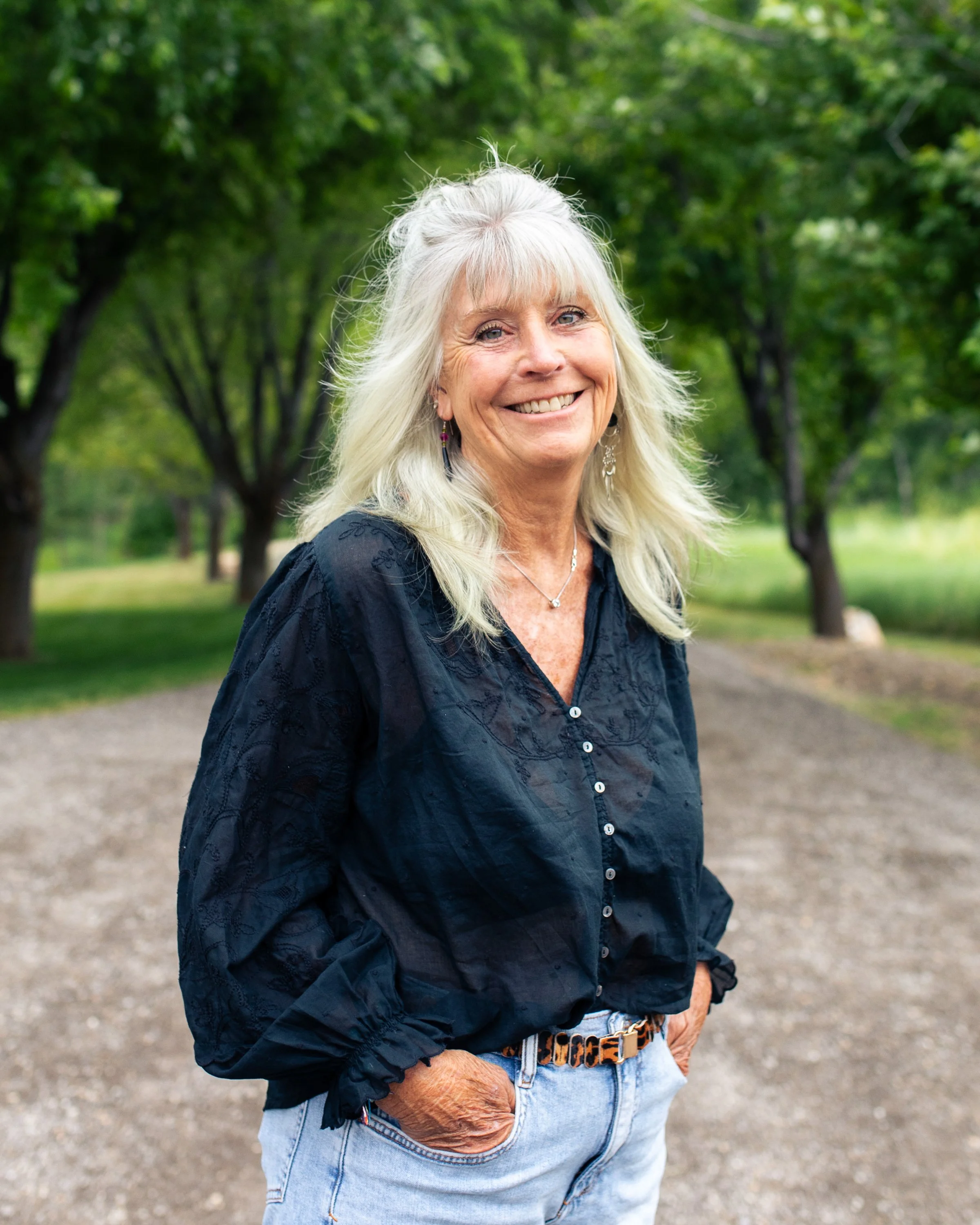 A smiling elderly woman with long white hair standing outdoors in a park with green trees in the background. She is wearing a navy blue blouse, light blue jeans, and jewelry including earrings and a necklace. She has her hands in her pockets.