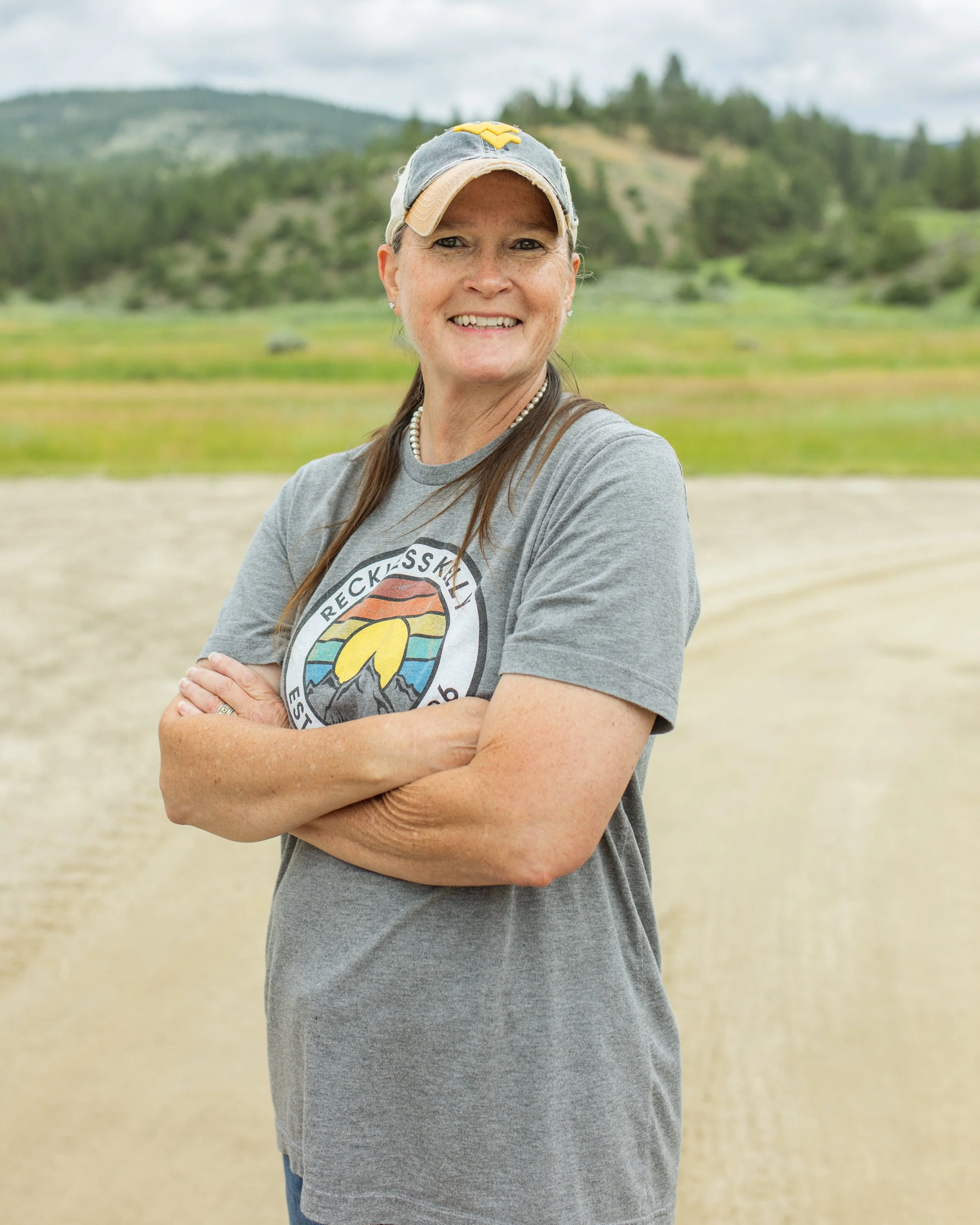A woman smiling outdoors in front of a landscape with green hills and cloudy sky, arms crossed, wearing a baseball cap and a gray T-shirt.