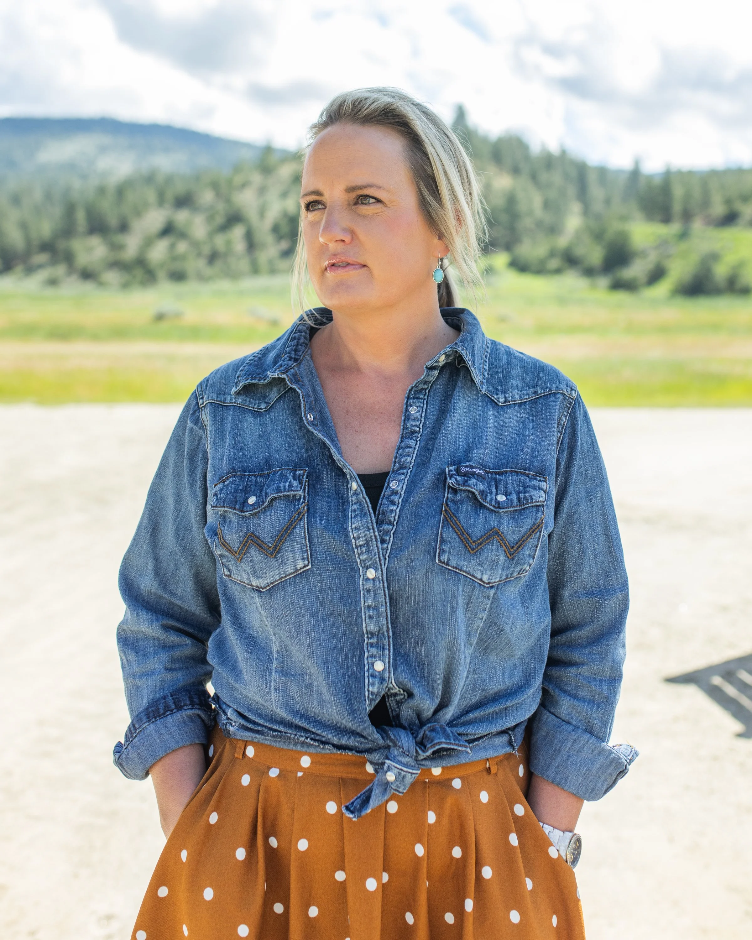 A woman with blonde hair wearing a denim jacket and a rust-colored polka dot skirt standing outdoors with a scenic background of trees and hills under a cloudy sky.