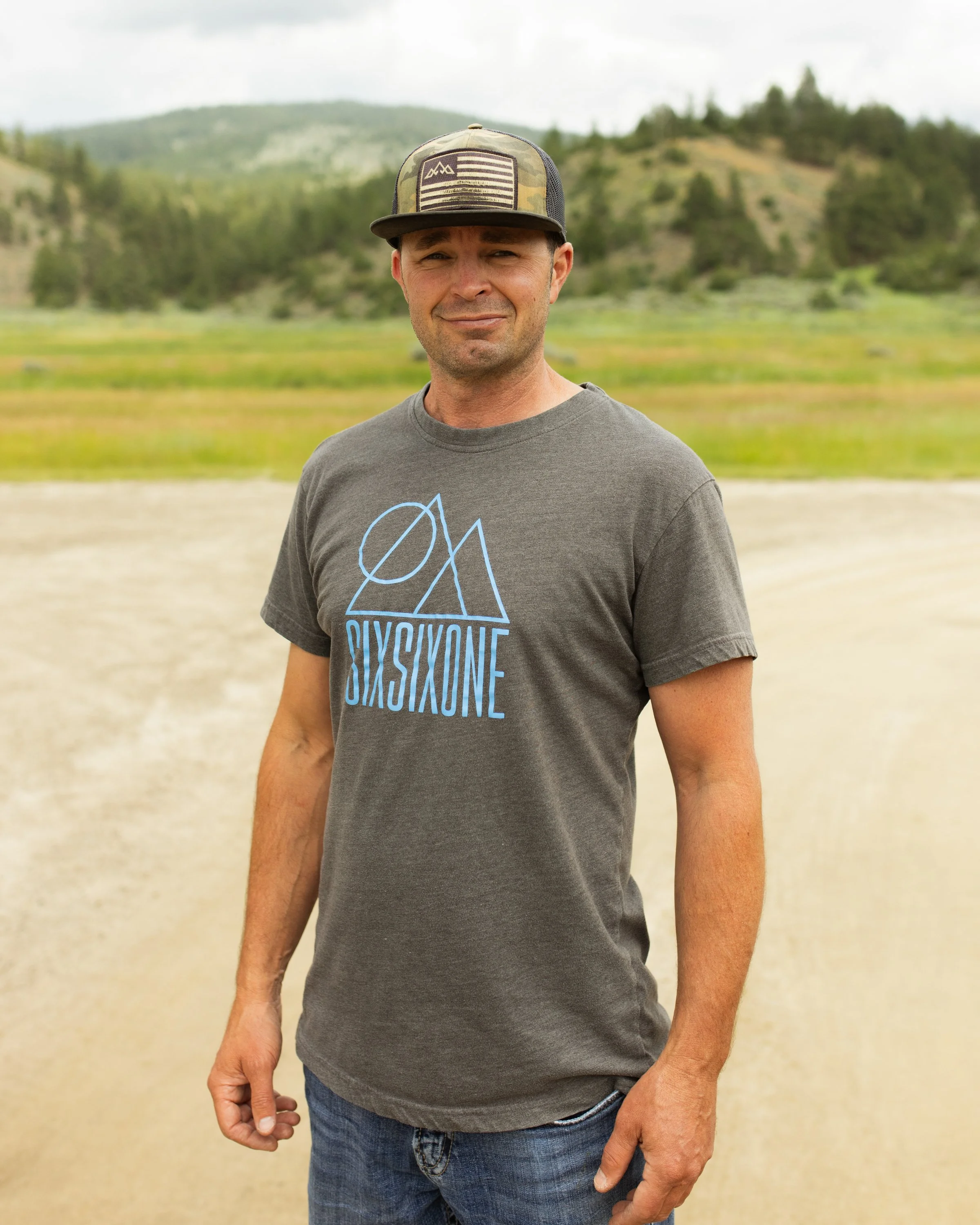 A man standing outdoors on a dirt road with hills and trees in the background, wearing a camouflage baseball cap and a gray T-shirt with a geometric design.