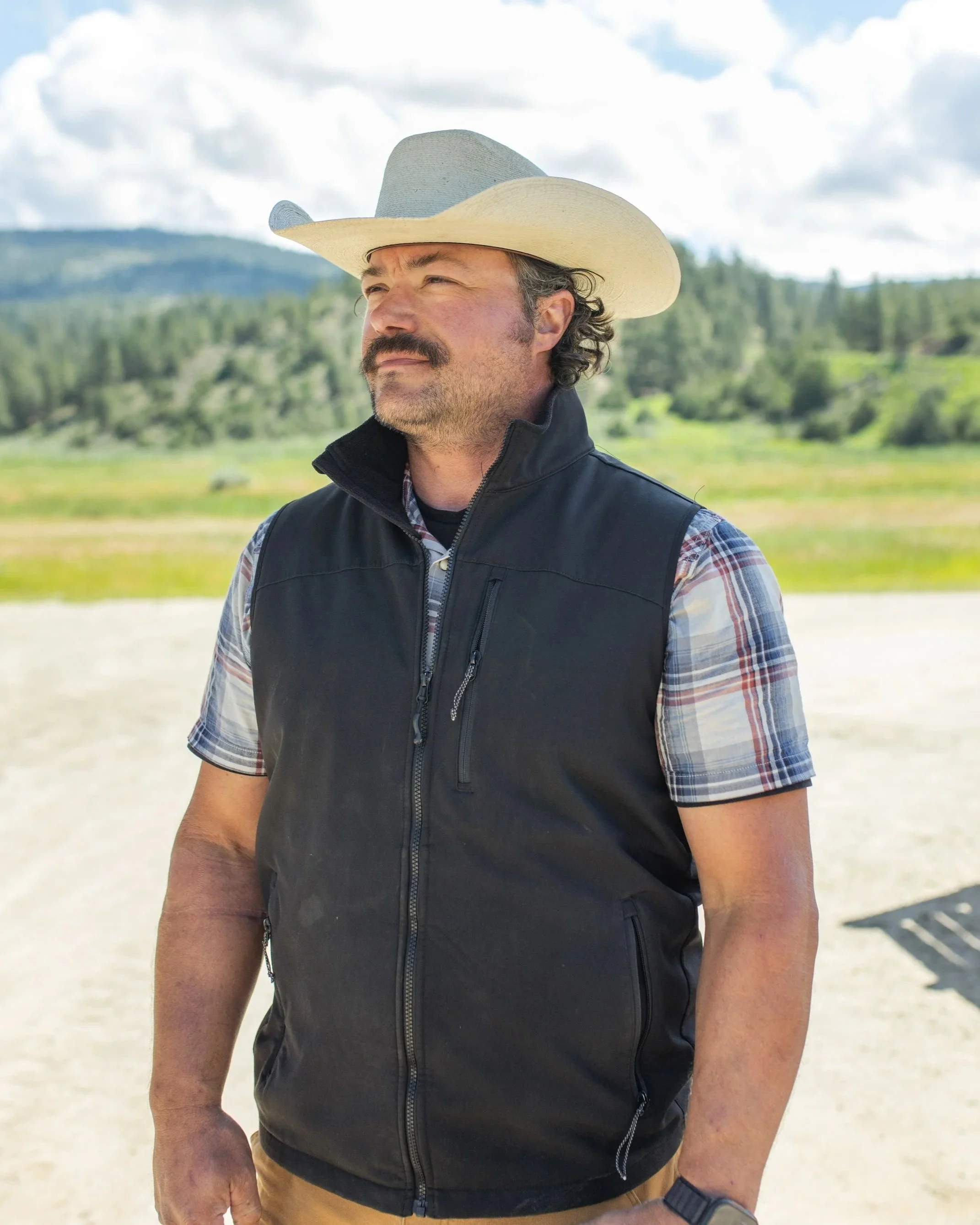 A man with a mustache and beard wearing a wide-brimmed cowboy hat, black vest, and plaid shirt outdoors in a rural area with mountains and trees in the background.