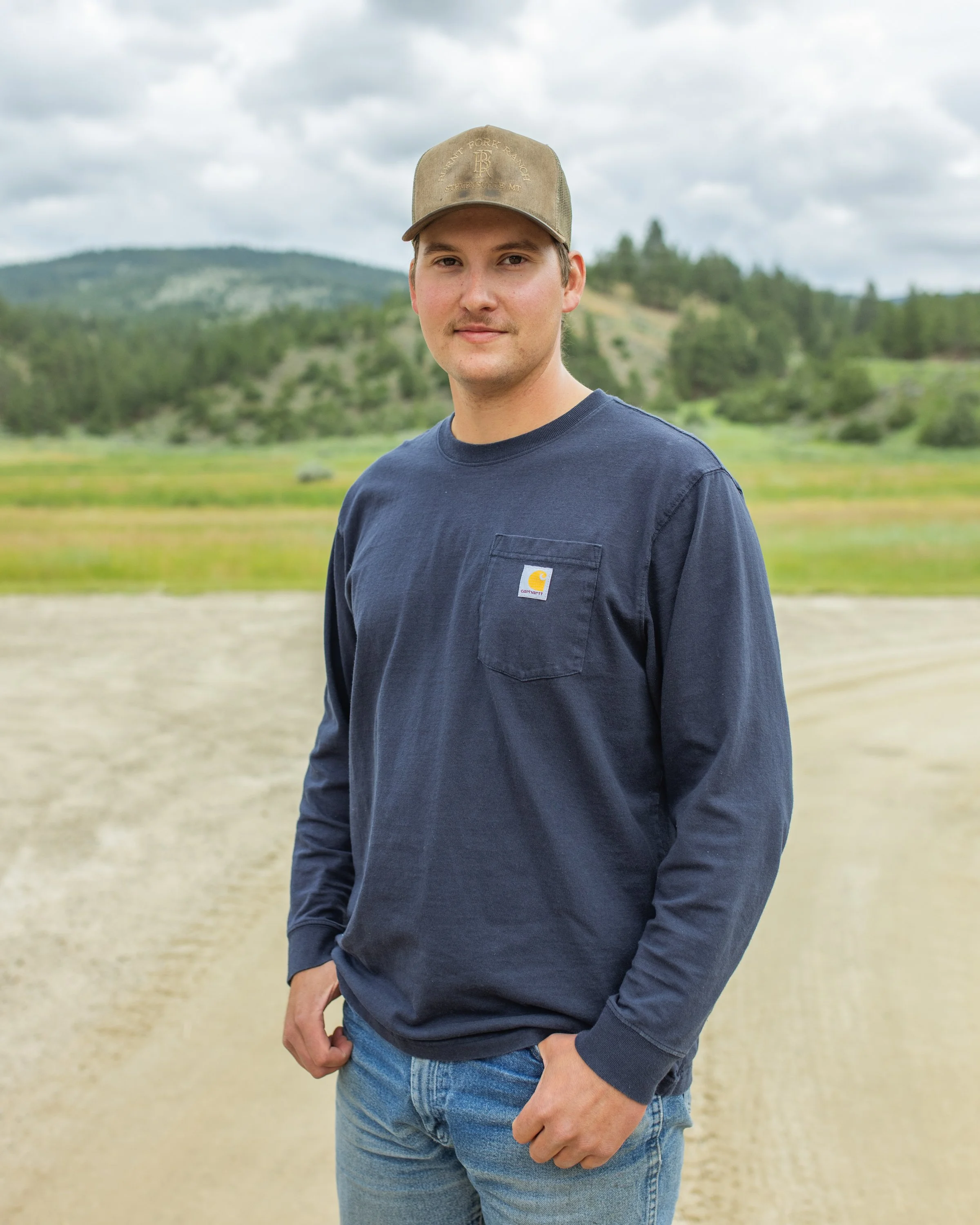 A young man with light skin wearing a beige baseball cap and a navy blue Carhartt long sleeve shirt, standing outdoors on a cloudy day in a grassy field with hills and trees in the background.