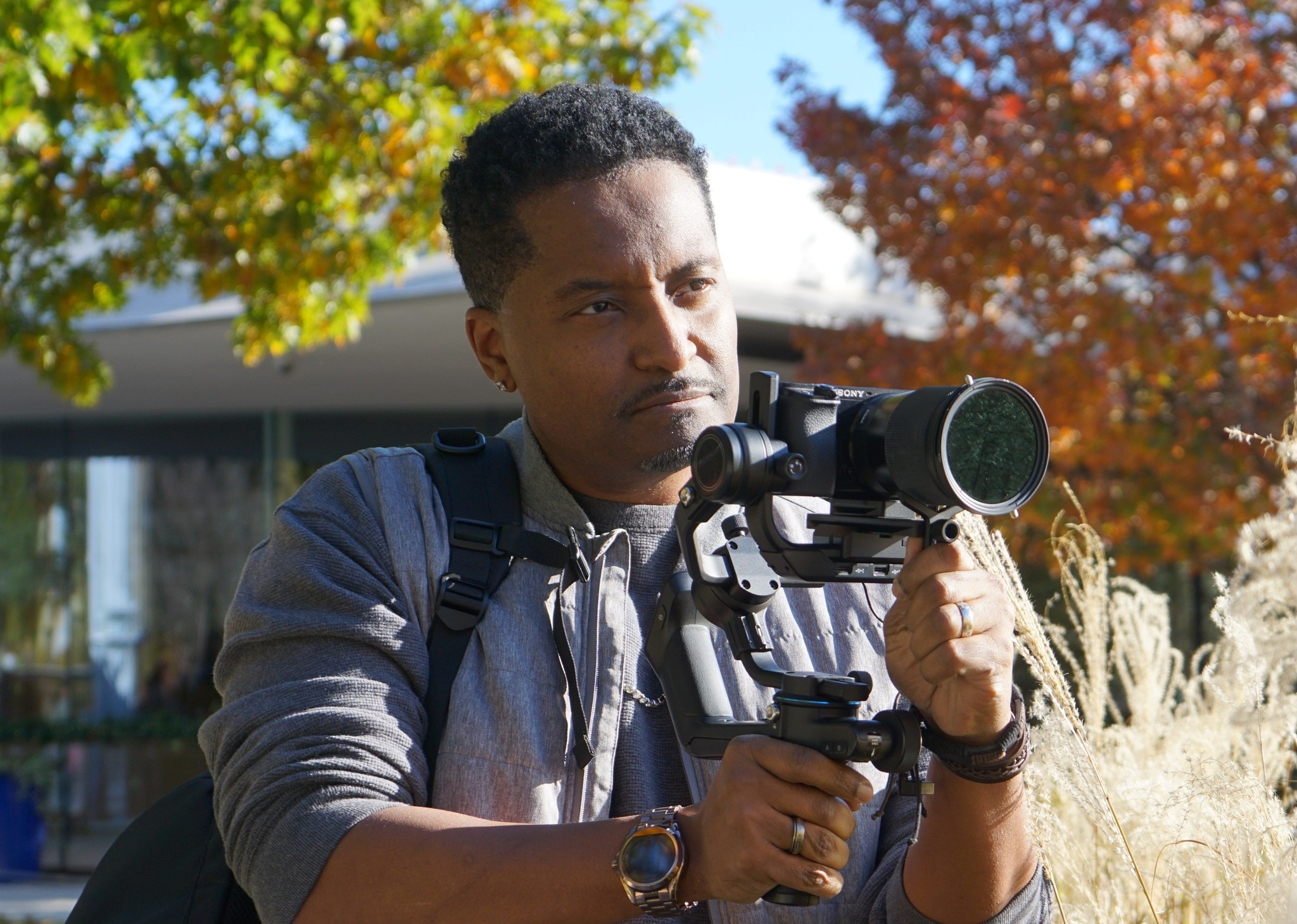 A man with short black hair and earrings is looking through a camera mounted on a tripod outdoors during autumn, with colorful trees in the background.