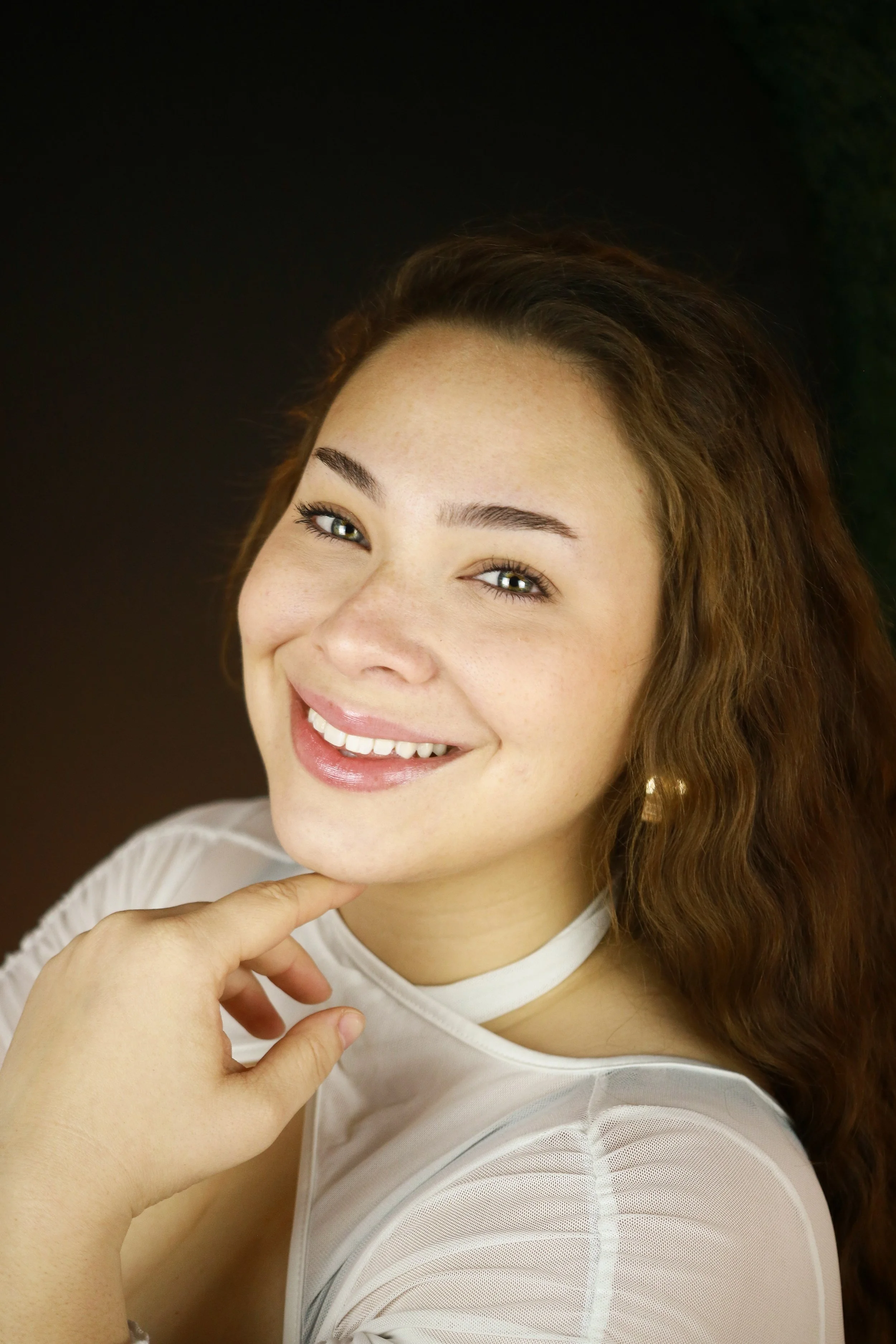 Close-up of a woman with brown hair, smiling, with her finger touching her chin, wearing a white top, in front of a dark background.