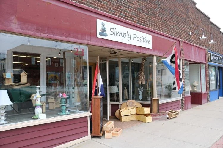 Storefront with a sign that reads "Simply Positive," showing various decorative items in the window and flags hanging outside.