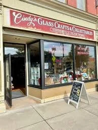 The storefront of Cindy's Glass Crafts & Collectables with large glass windows, a red sign, and a sidewalk chalkboard sign outside.