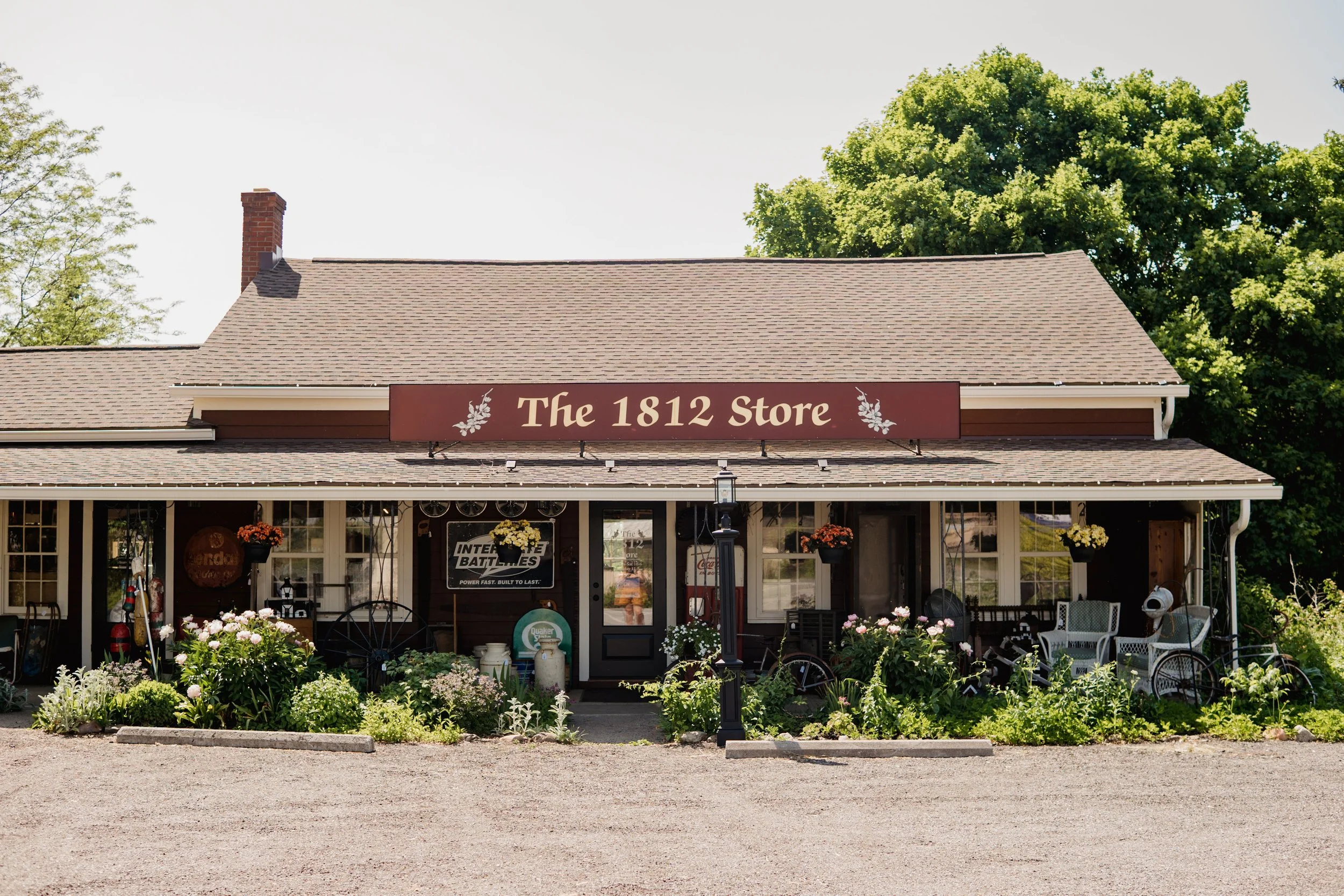 Front view of a rustic store called 'The 1812 Store' with a maroon sign, surrounded by greenery and flowers, featuring outdoor seating, vintage decor, and a gravel parking area.
