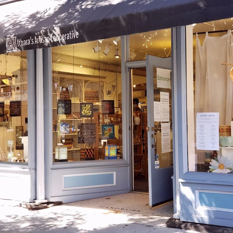 The entrance of a shop called Ithaca's Artisan Cooperative with large glass windows displaying artwork, and a sign on the door labeled "Entrance." A person is visible inside the shop.