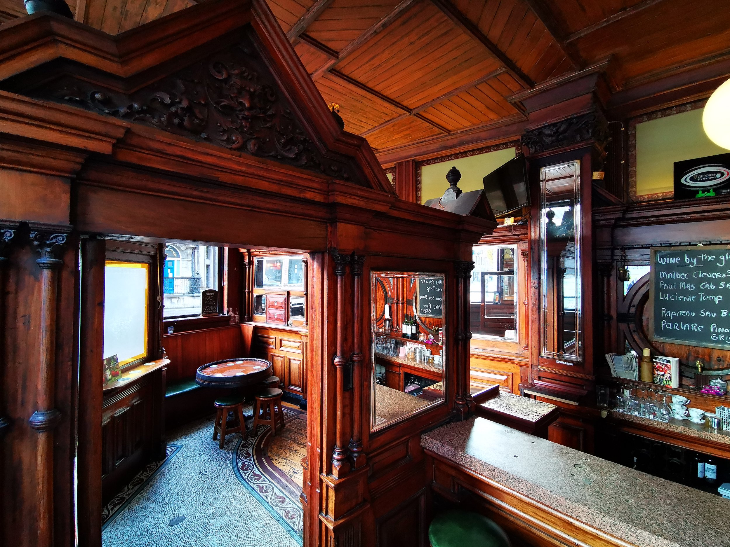 Inside a cozy bar with dark wood paneling, a small round table with stools, windows letting in natural light, and a chalkboard menu listing wines.