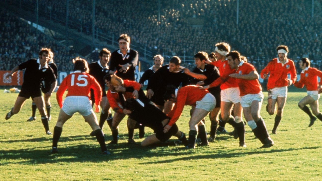 A rugby match with players in black and red uniforms engaging in a tackle on the field during daytime.