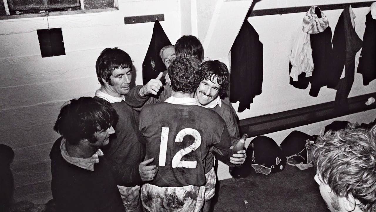 A group of men, some with dark hair and some with curly hair, are celebrating together. One man with curly hair is smiling and holding a bottle, wearing a sports jersey with the number 12. The others are gathered around him, in what appears to be a l