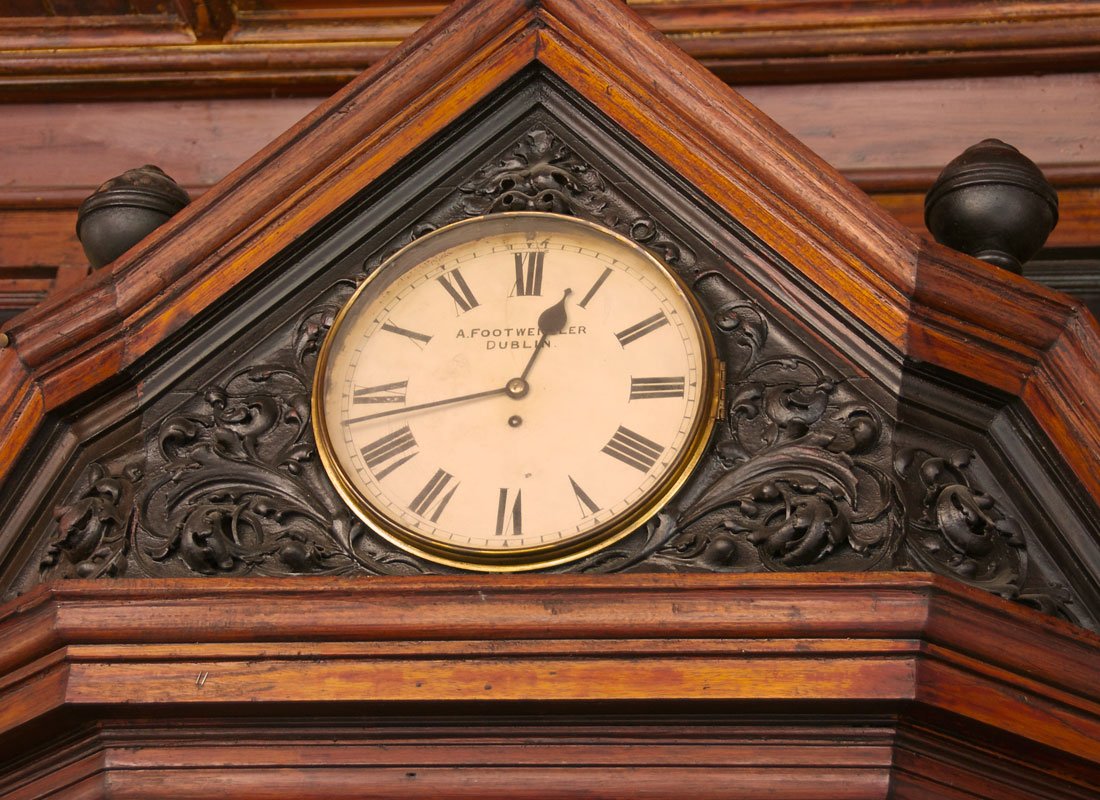 A vintage wall clock with Roman numerals enclosed in ornate black wood carving and a wooden frame, showing the time as 12:03, labeled 'A. Footwer Dublin'.