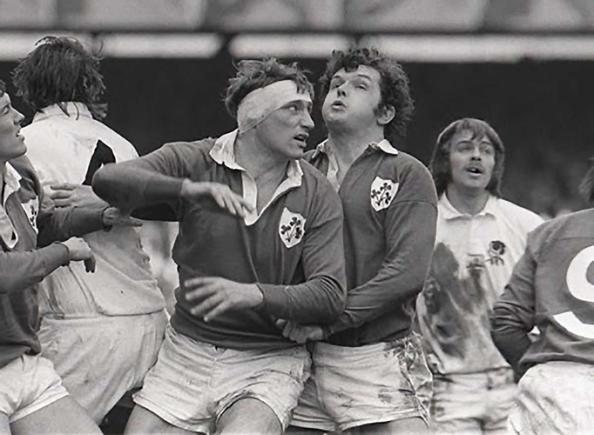 Black and white photo of rugby players on the field, with two players embracing and others around them, some wearing rugby jerseys with crests, celebrating a moment during a game.