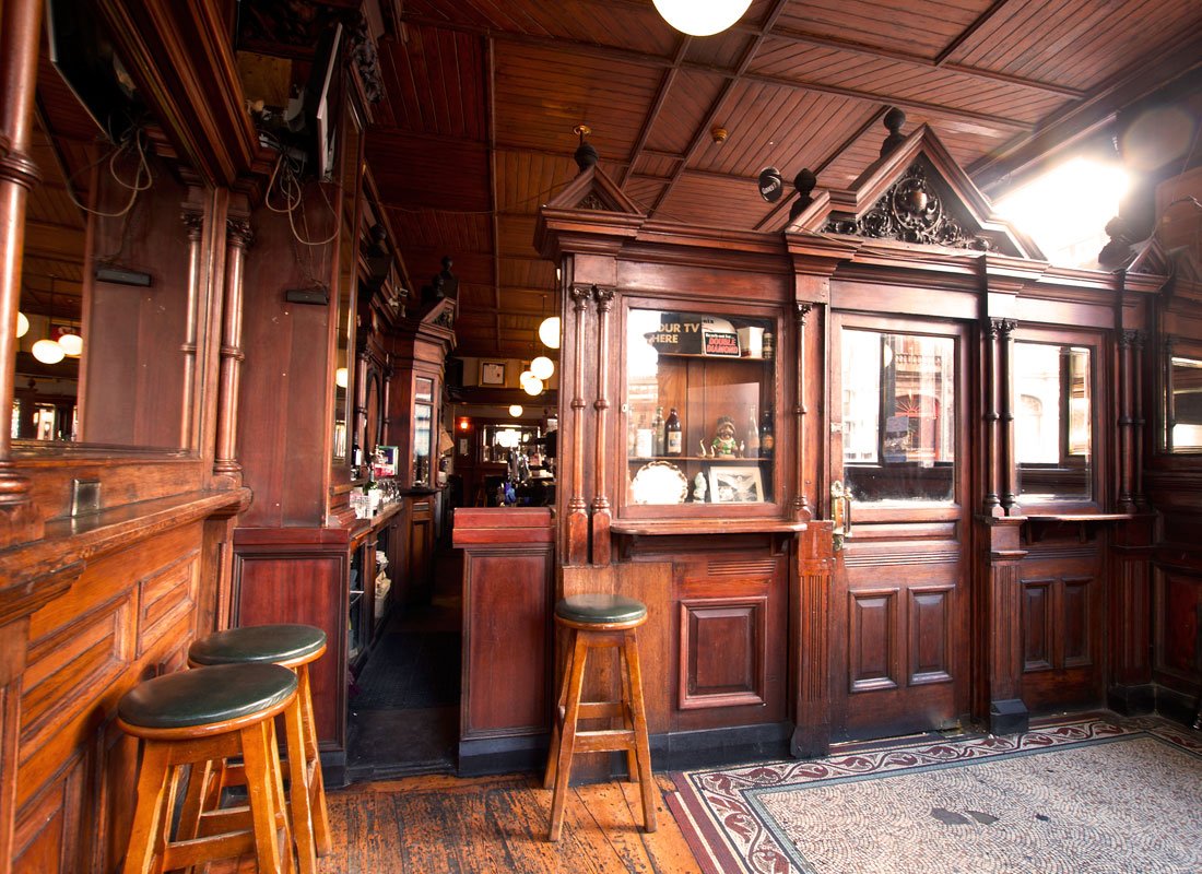 Interior of a bar with dark wood paneling, a small windowed partition, and wooden stools with green cushions in a cozy pub setting.