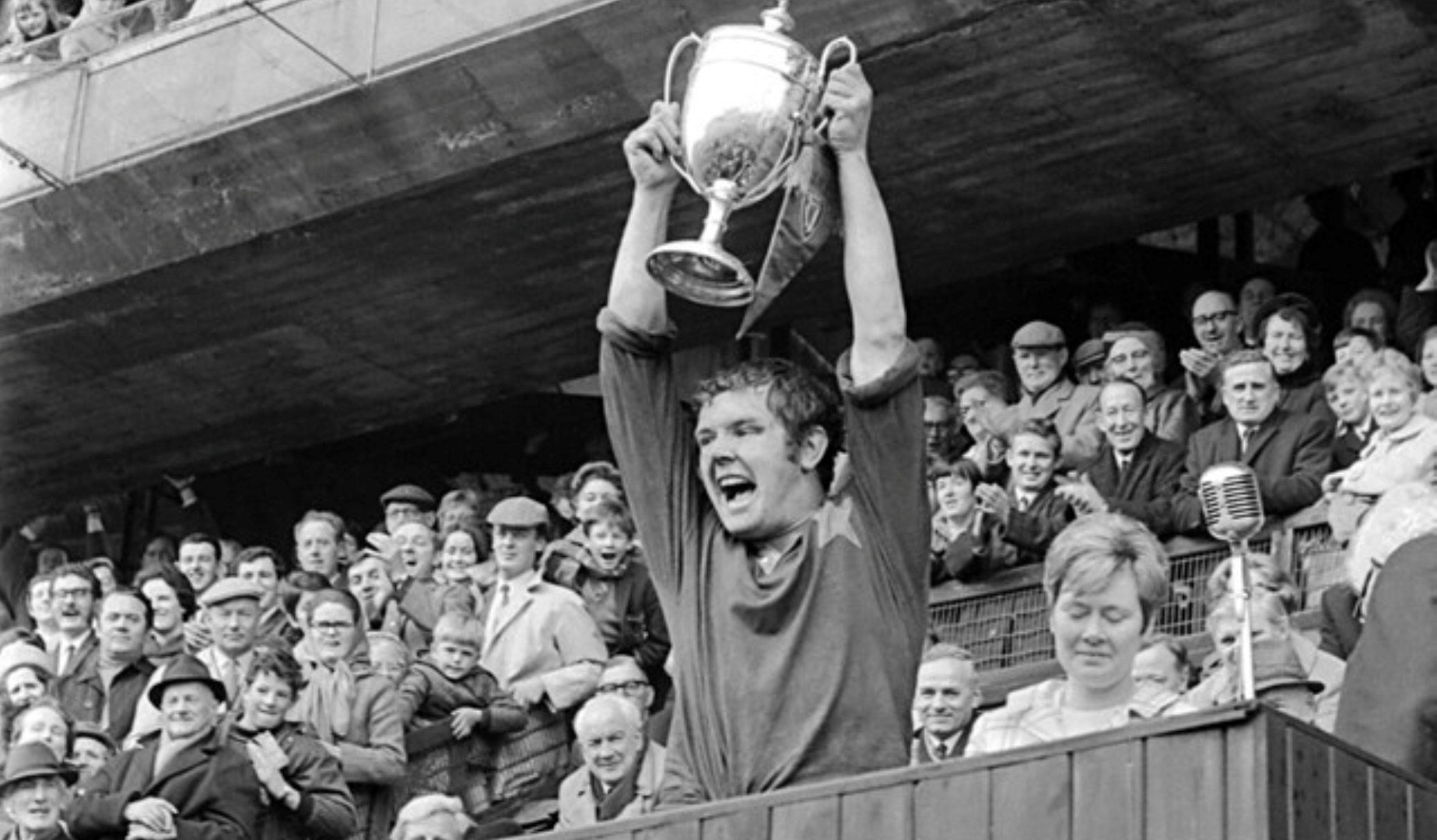 Black and white photo of a young man in a sports jersey celebrating, holding a trophy above his head, surrounded by a cheering crowd in a stadium.