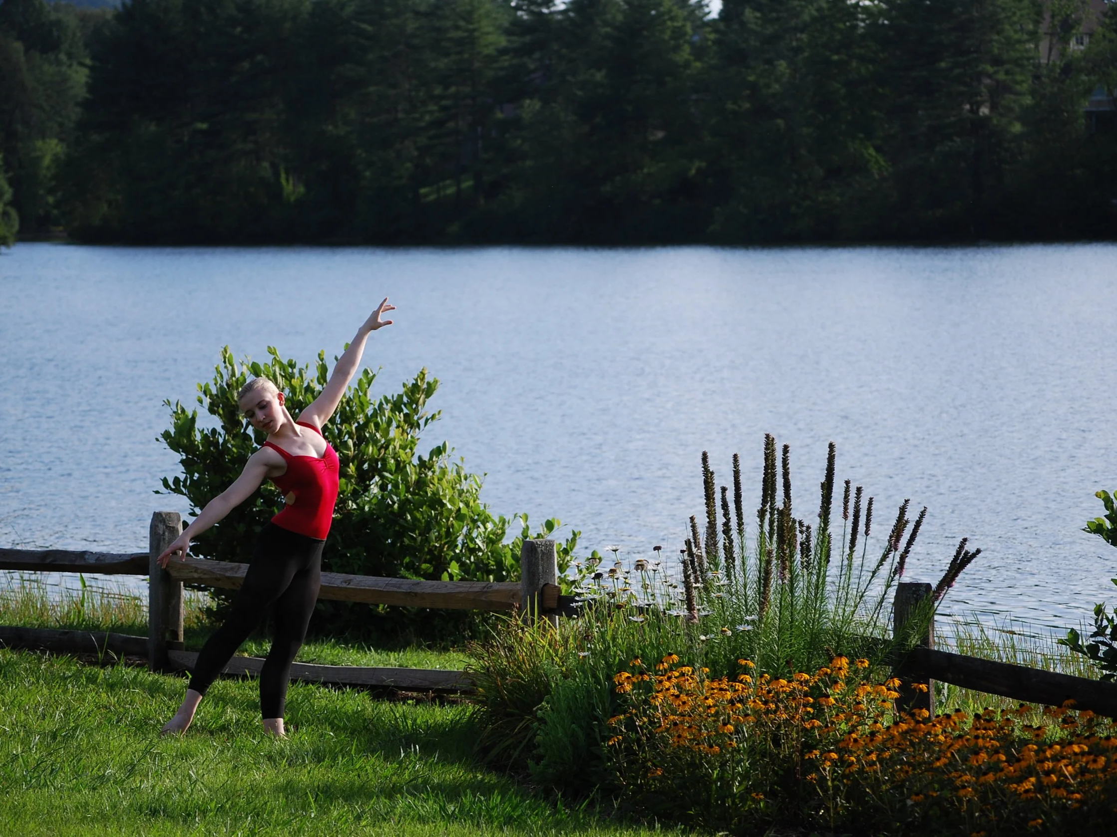 A dancer in a red leotard posed in a reflective manner in front of lake with beautiful foliage around.