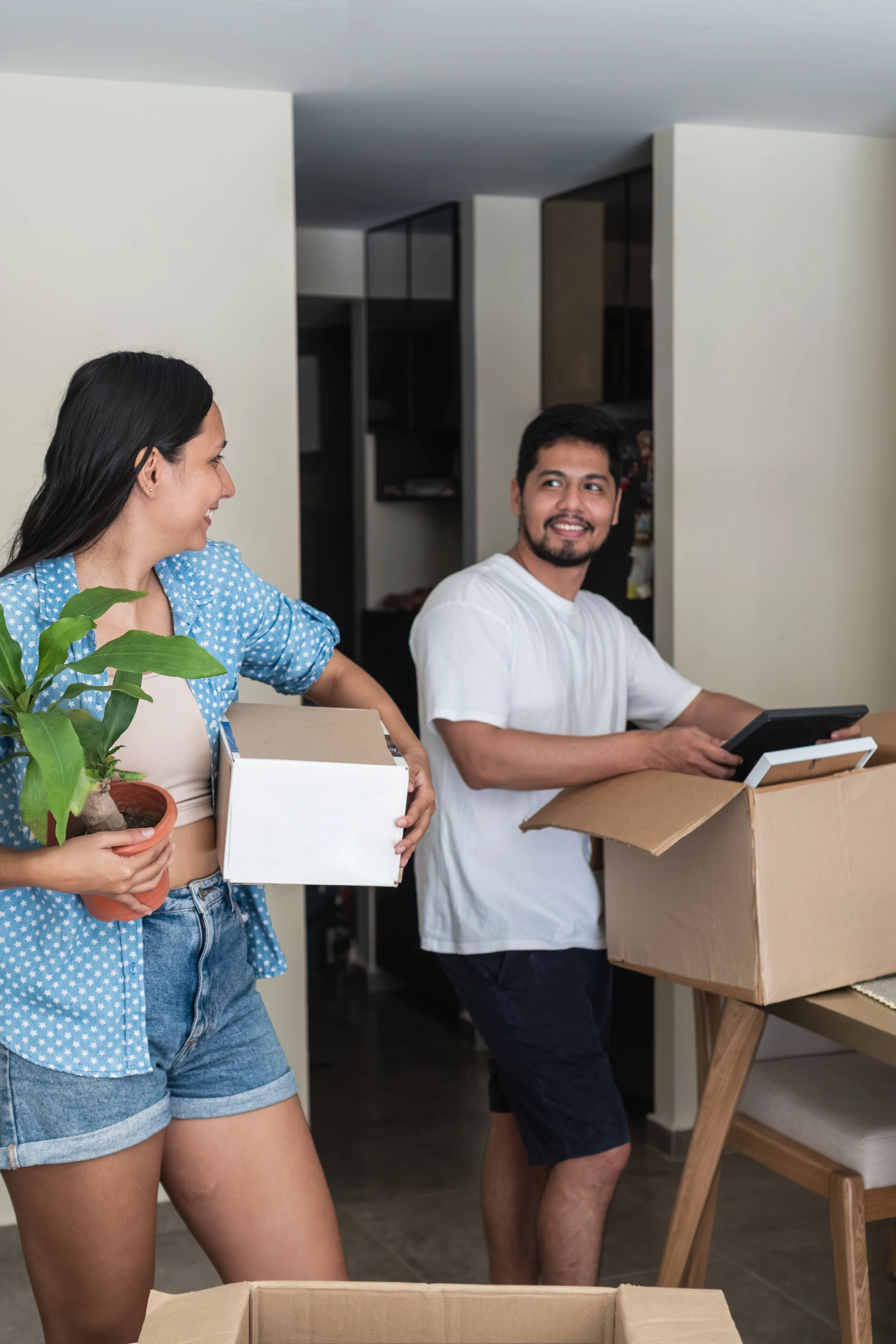 A woman in a blue polka dot top and denim shorts holding a potted plant and a white box, smiling at a man in a white t-shirt and dark shorts who is carrying a cardboard box. They are inside a house with a wooden table and kitchen cabinets in the background.