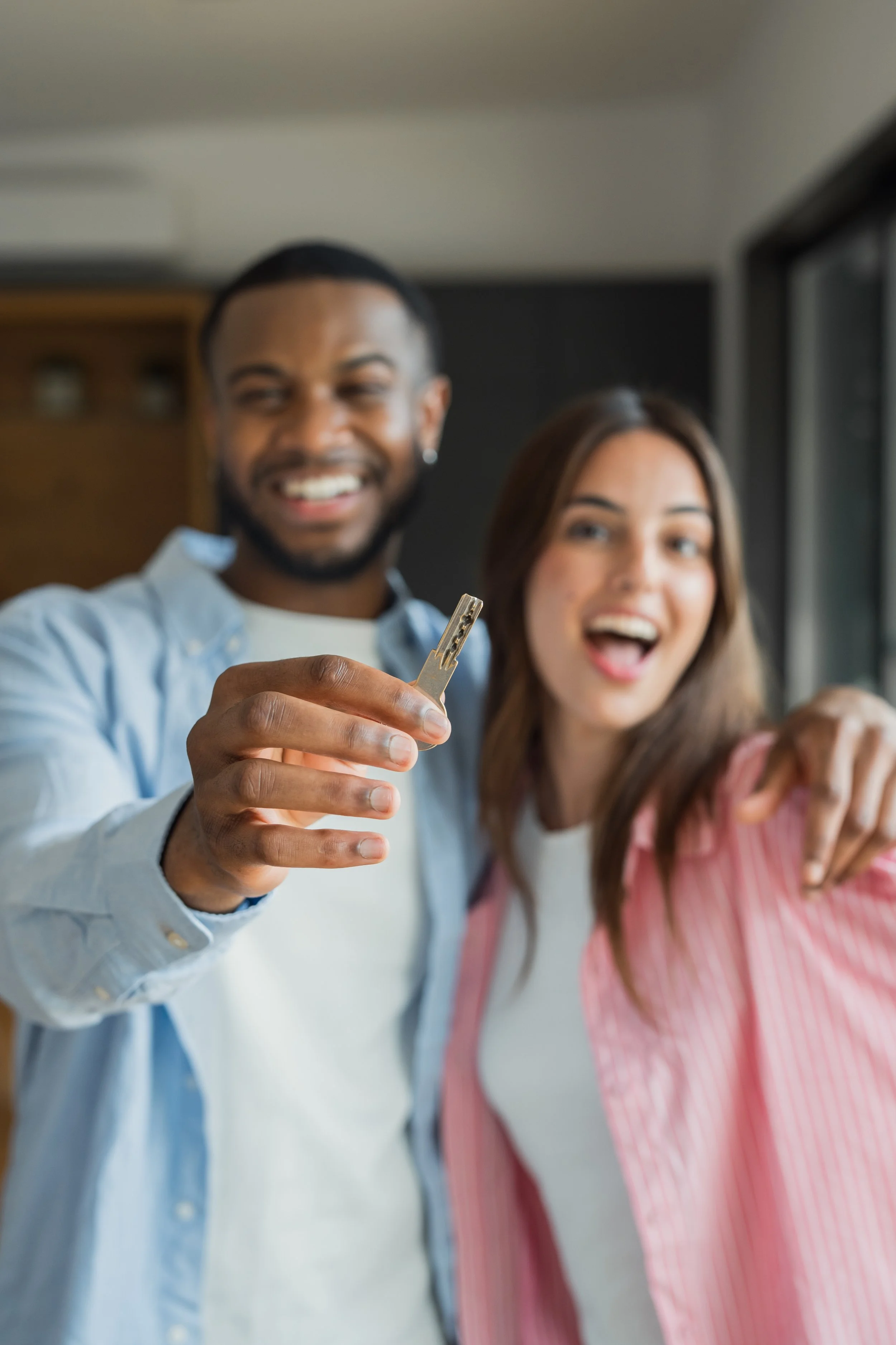 Two smiling people, a man and a woman, taking a selfie together indoors. The man is holding the camera, and the woman is pointing towards the camera.