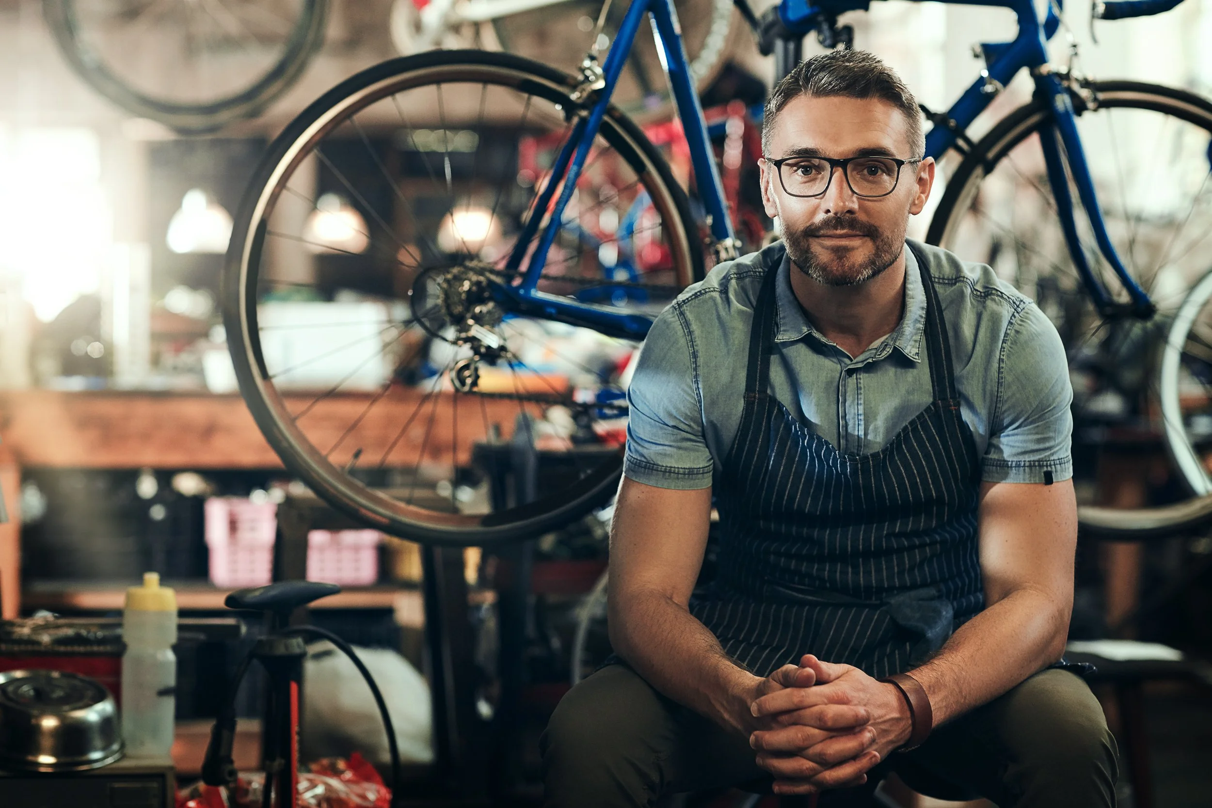 A man with glasses and a beard, wearing a denim shirt and apron, sitting inside a bicycle workshop with bikes hanging on the wall behind him.