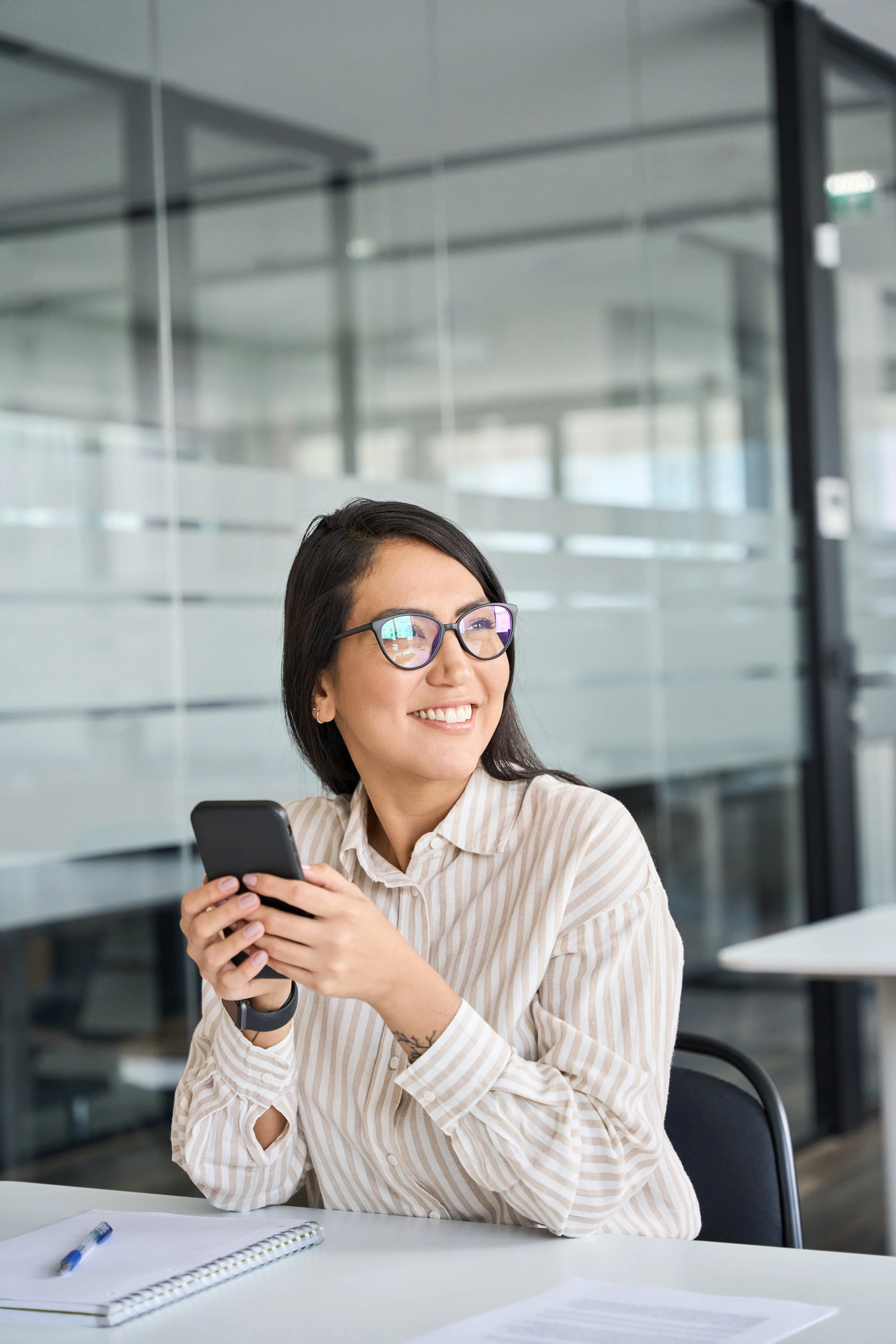 A woman with dark hair, glasses, and a striped beige button-up shirt sitting at a table in an office, smiling while looking at her smartphone, with notebooks and a pen on the table and glass walls in the background.