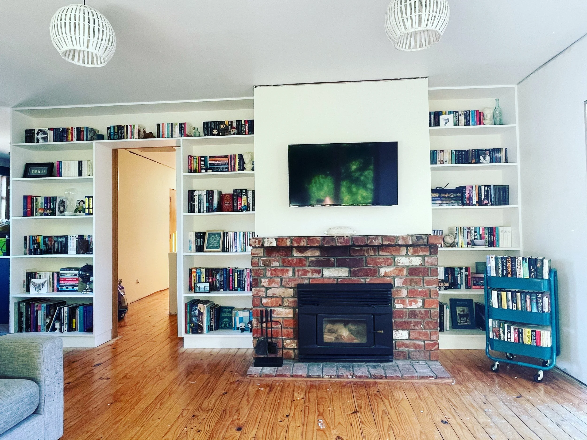 Living room with brick fireplace, mounted TV, white bookshelf filled with books, and hardwood floors.