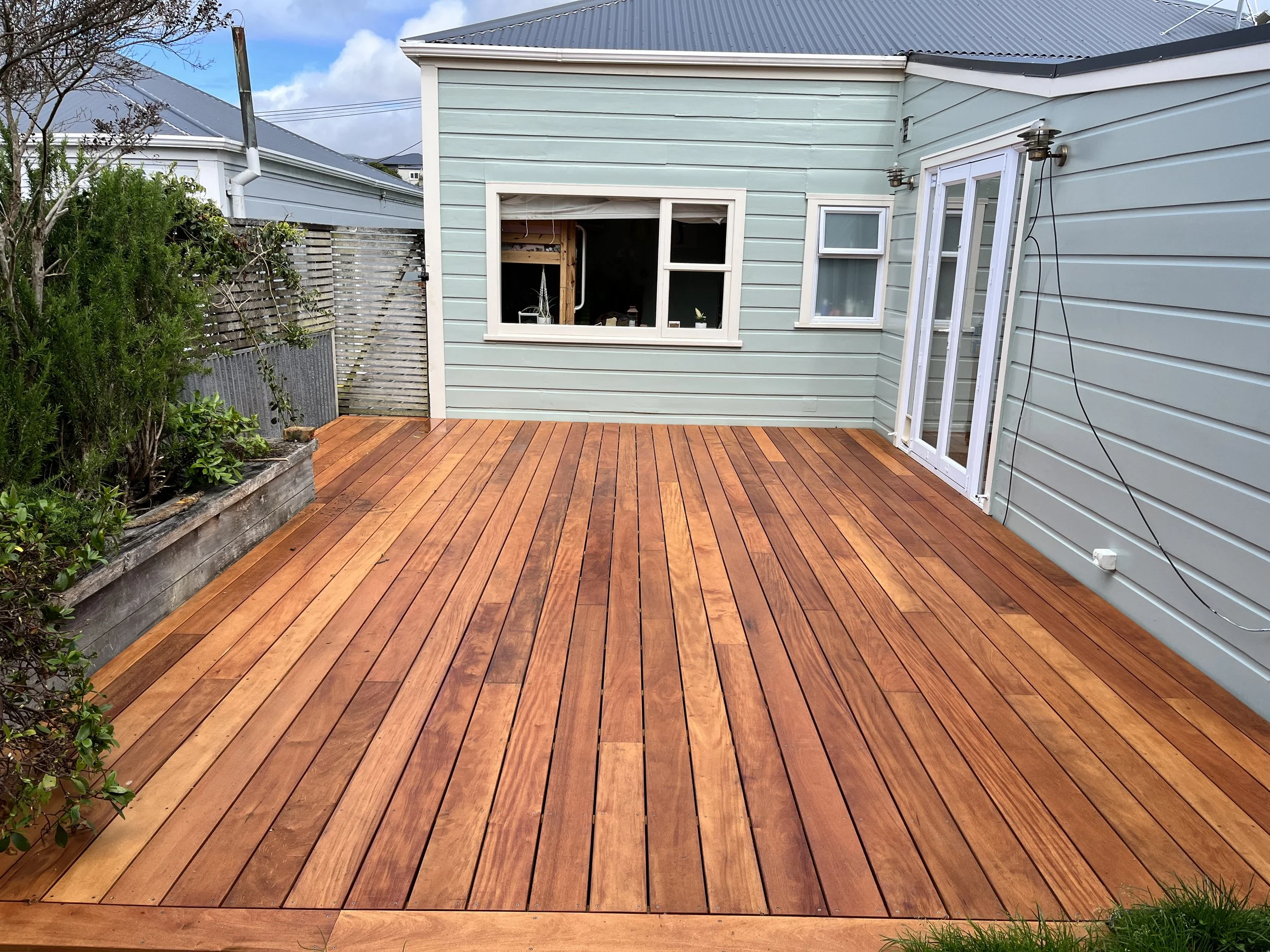 Clean and newly finished wooden deck attached to a light blue house with sliding glass doors and windows, with some greenery and garden bed on the side.