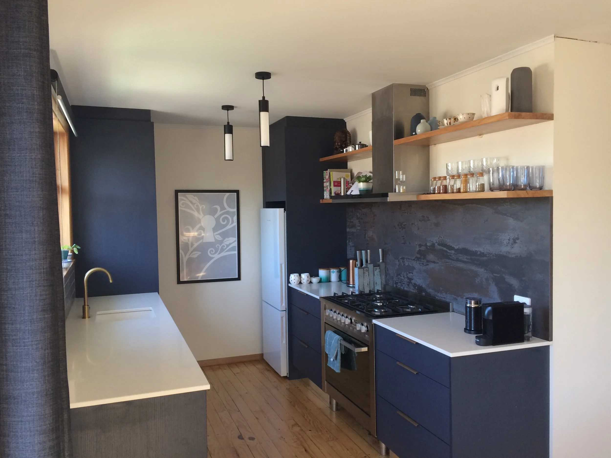 Modern kitchen with dark blue cabinets, white countertop, open wooden shelves, and a gray textured backsplash. Contains a gas stove, coffee maker, and glass jars along the shelves. Light wood flooring and a framed art piece on the wall.