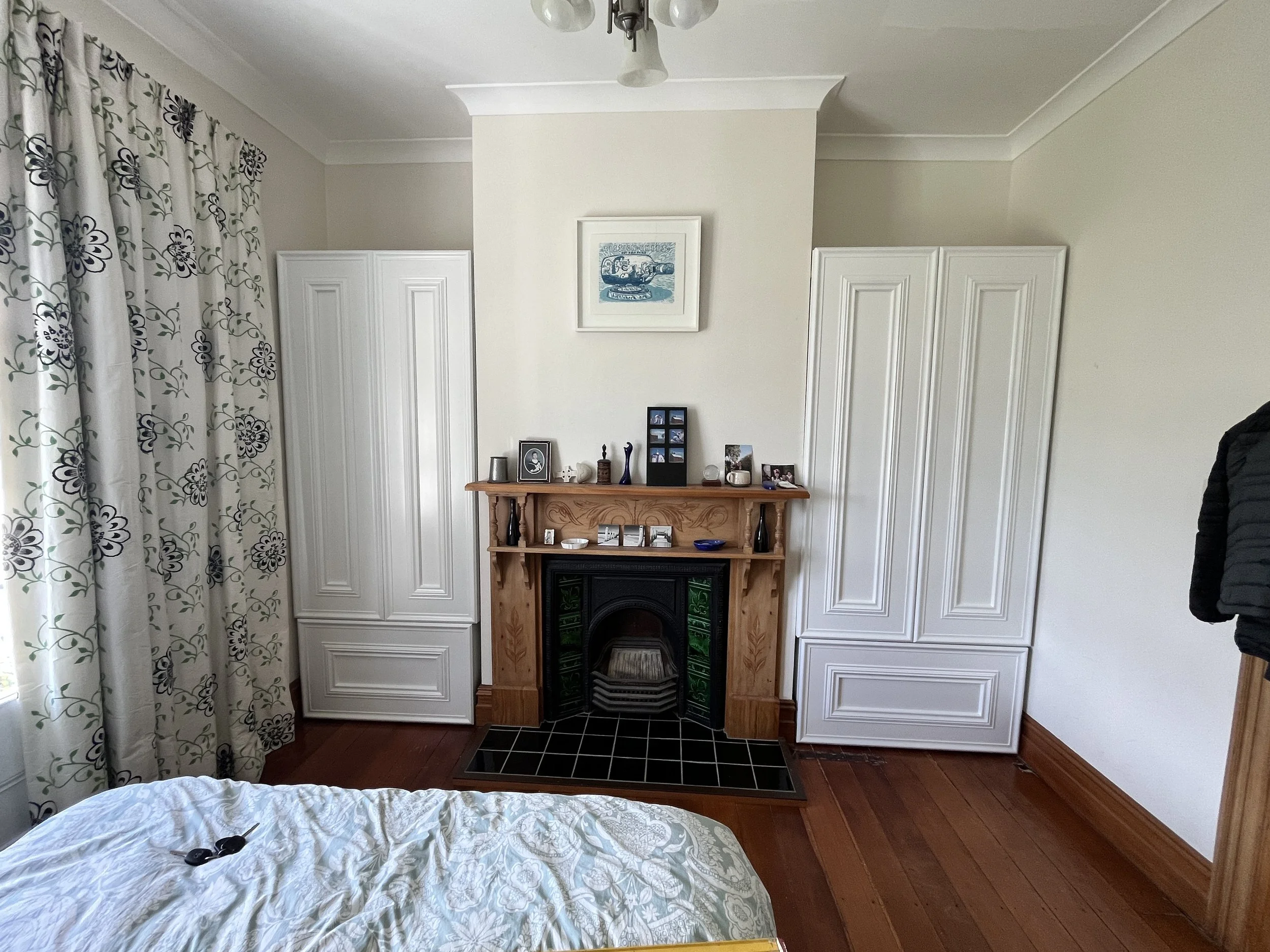 A bedroom with a fireplace surrounded by two tall white cabinets, a wooden mantel with framed photos and decorative items, a bed with white bedding, patterned curtains, and wooden floors.