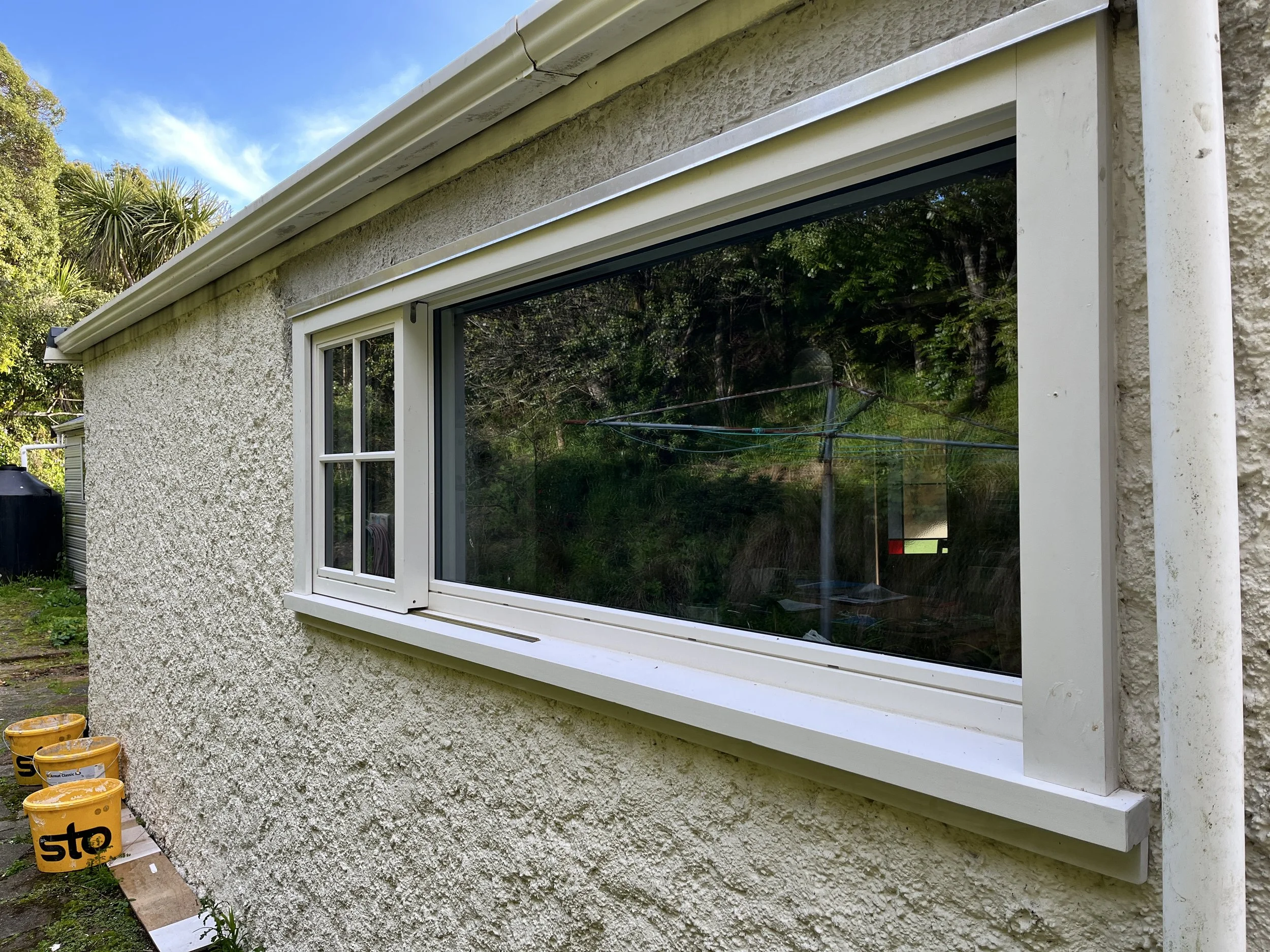 Outside wall of a house with a large rectangular window, textured stucco wall, three yellow buckets on the ground, and greenery reflected in the window.