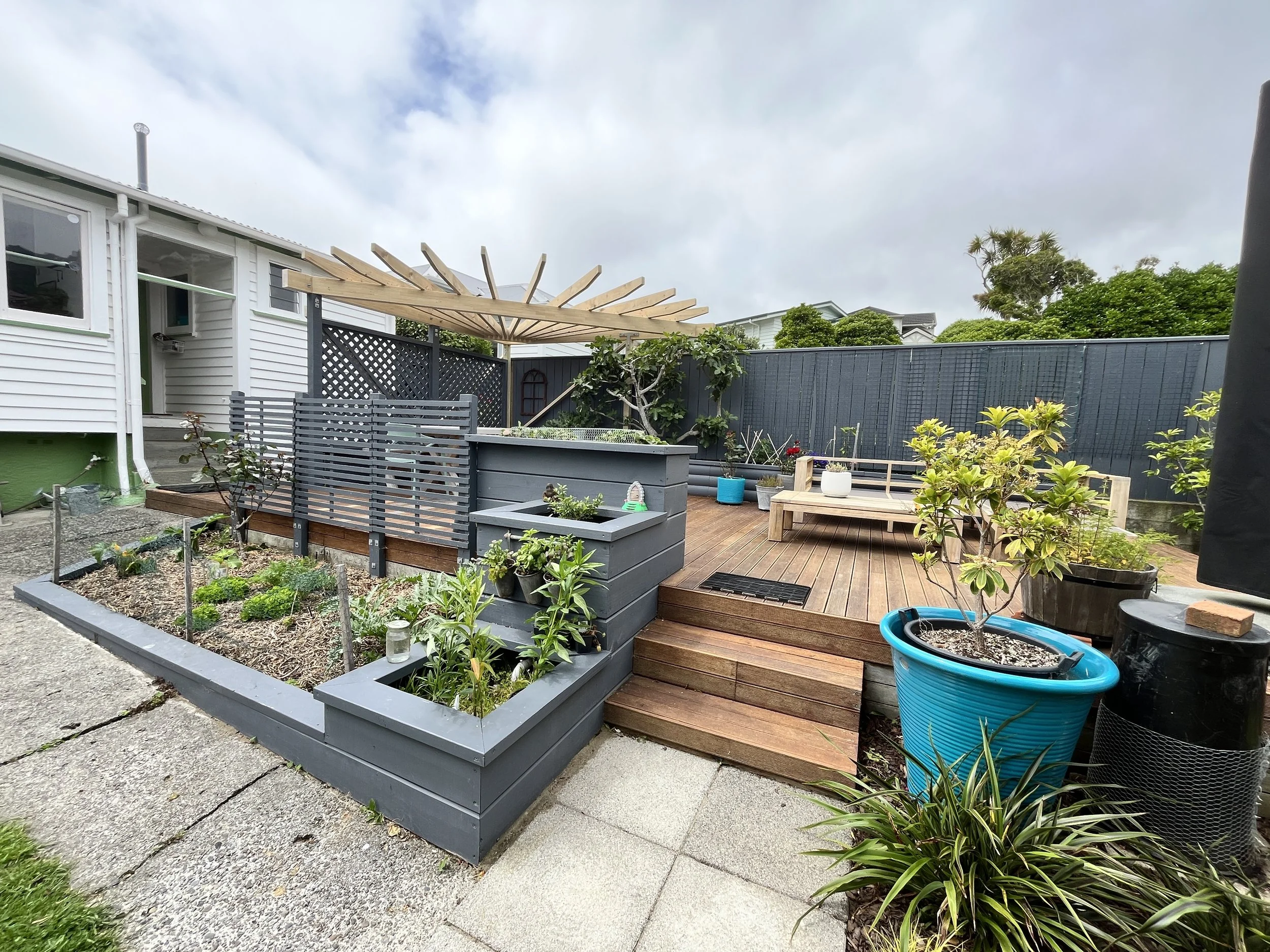 Backyard patio with raised garden beds, potted plants, wooden deck, outdoor seating, and a pergola with a cloudy sky. Karori wellington