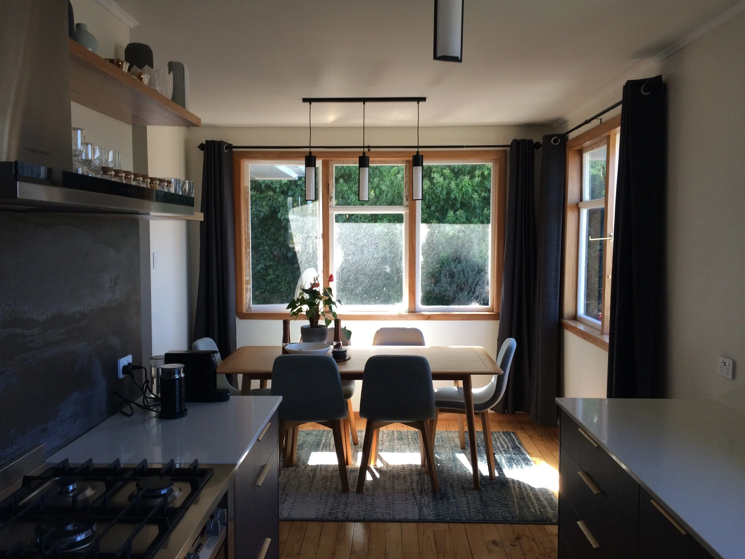 Kitchen and dining area with wooden table, six chairs, large windows with black curtains, and a view of green bushes outside.