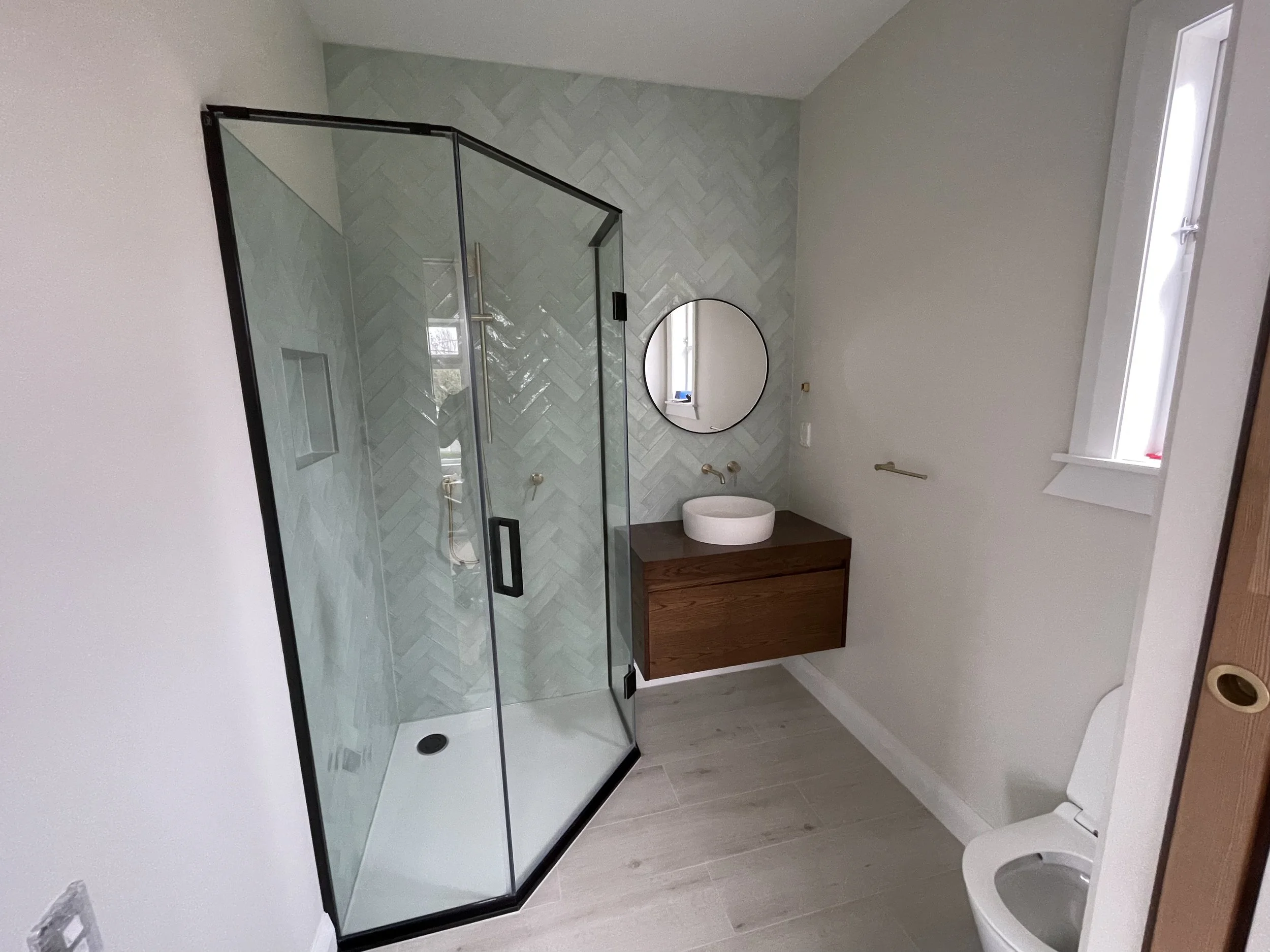 Modern bathroom with a glass-enclosed shower, round mirror, wooden vanity with white vessel sink, and a toilet near a window.