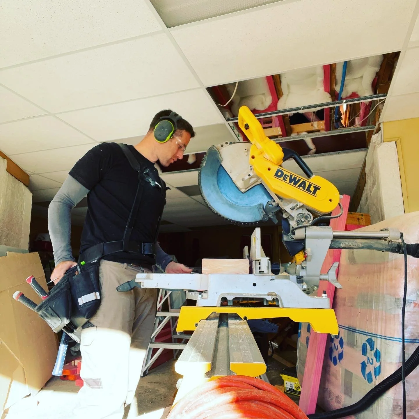 A man wearing hearing protection and a tool belt operates a yellow DeWalt miter saw cutting a piece of wood in a workshop with an unfinished ceiling.
