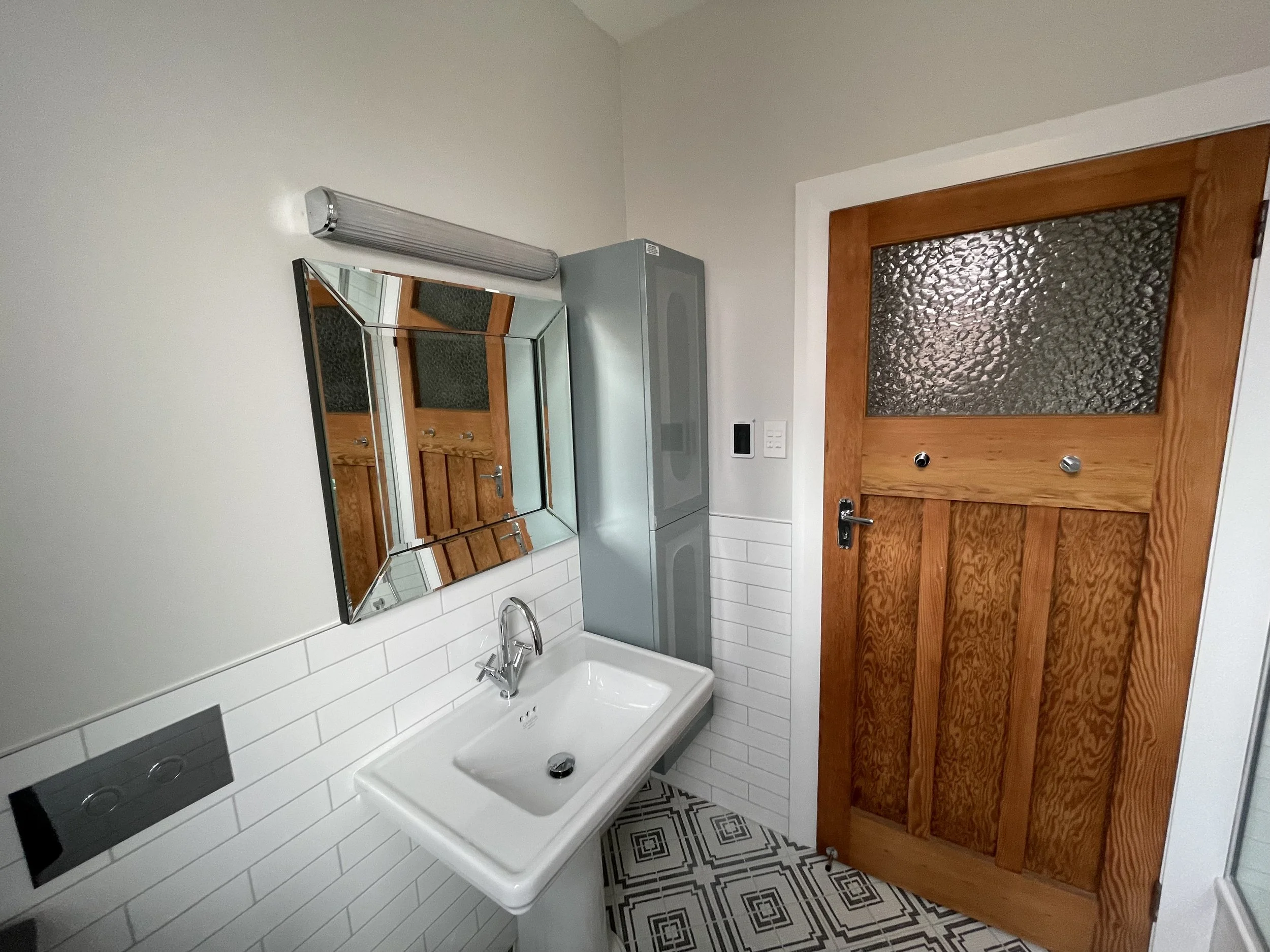 Bathroom with a white sink, a mirror, a wooden door with a textured glass window, white tiled walls, and patterned tile floor.