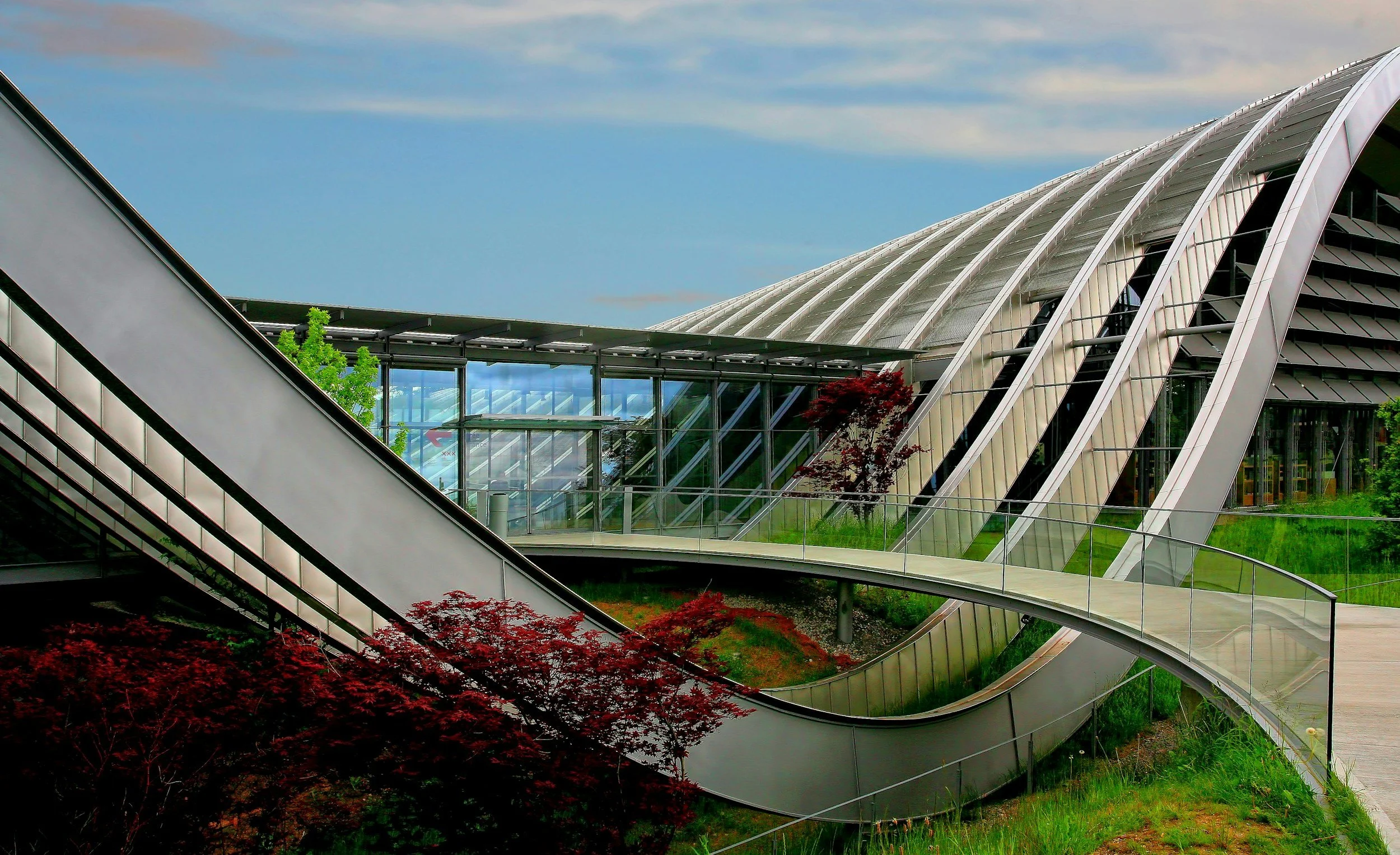 Modern building with curved glass and metal architecture, surrounded by green grass, trees, and a blue sky.