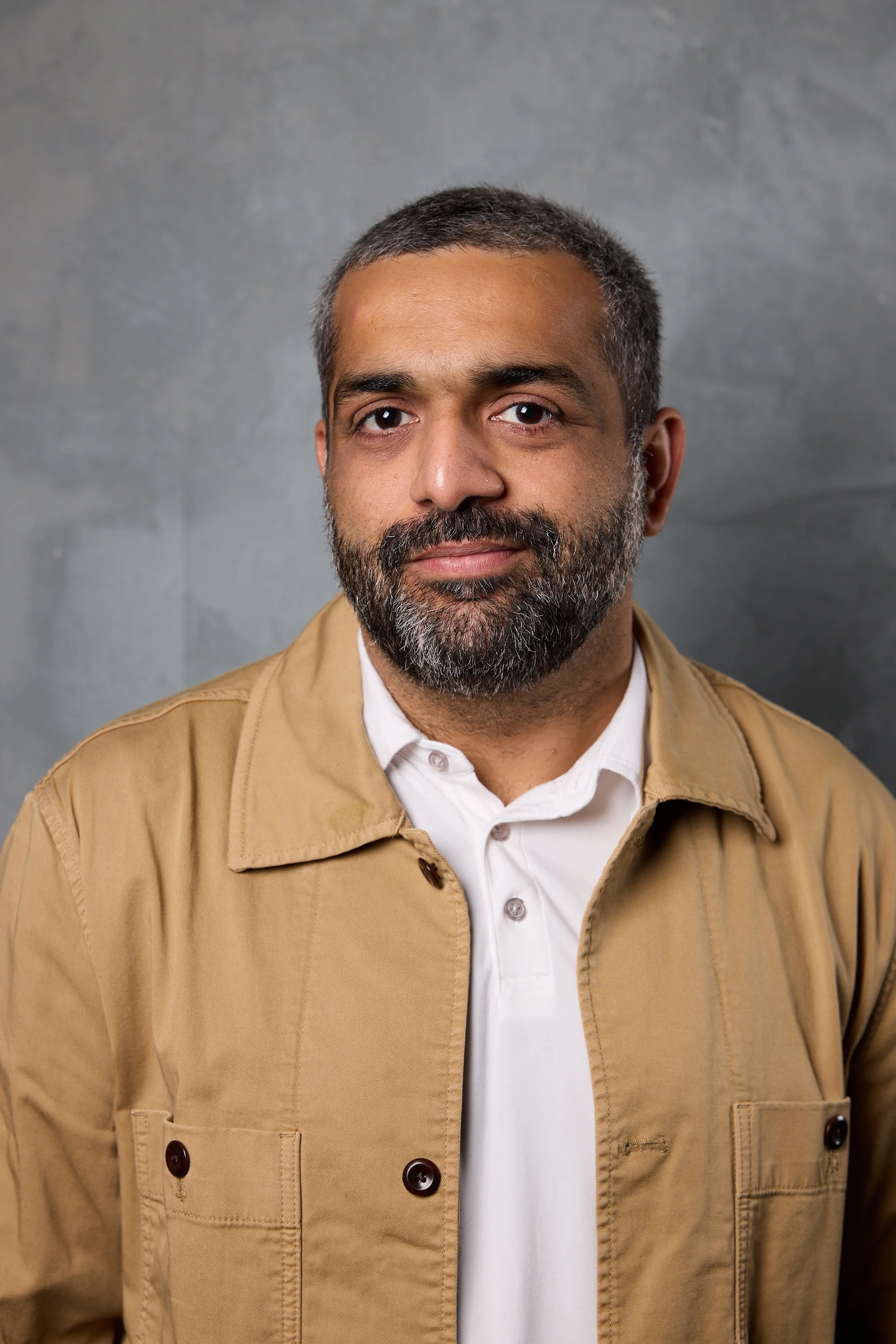 Portrait of our advisor, Kanav Hasija, a man with short dark hair and a beard wearing a tan jacket over a white shirt on a gray background.