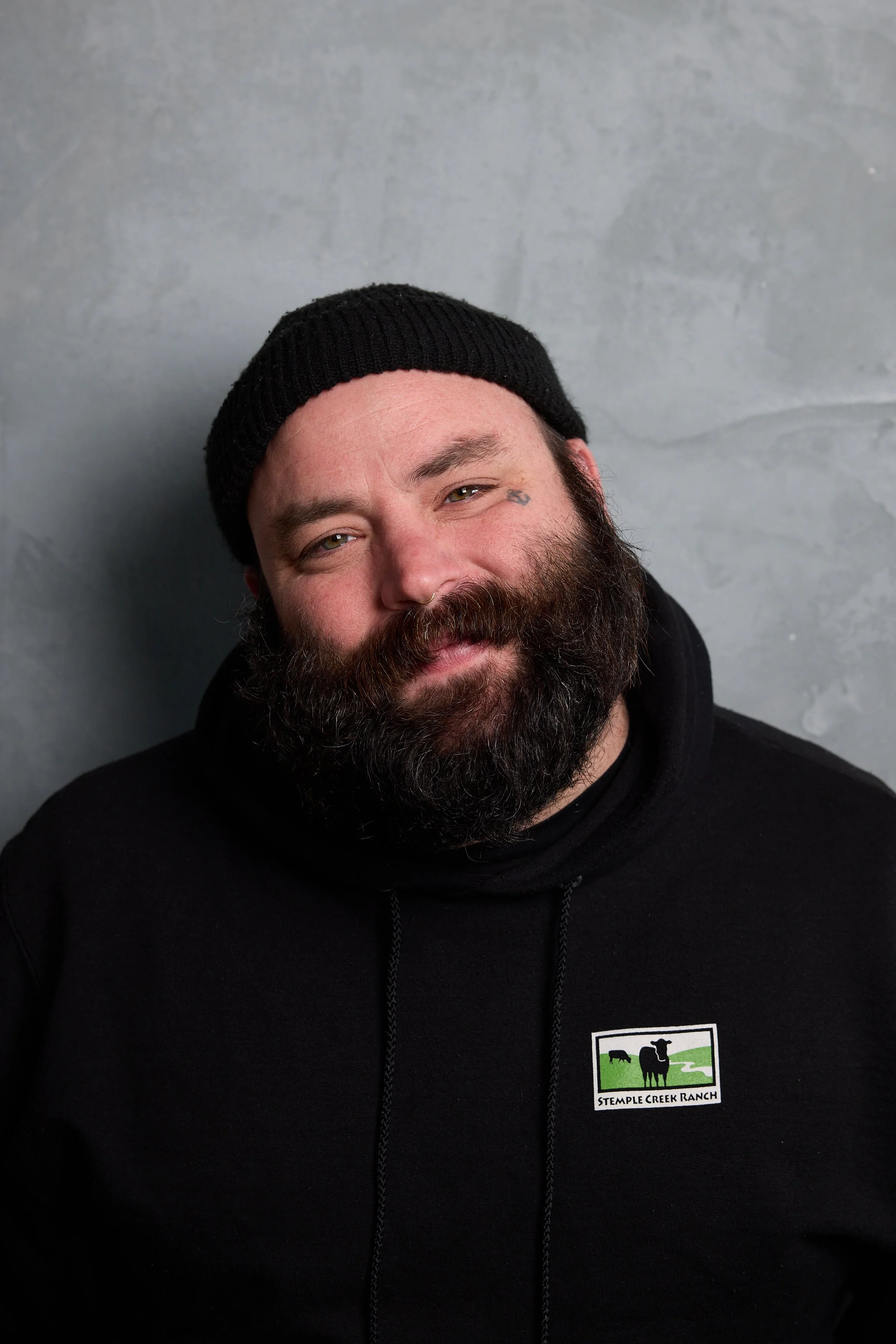 Portrait of our co-founder, Seth Stowaway, a man with a beard and mustache, wearing a black beanie and black hoodie, against a gray textured background.